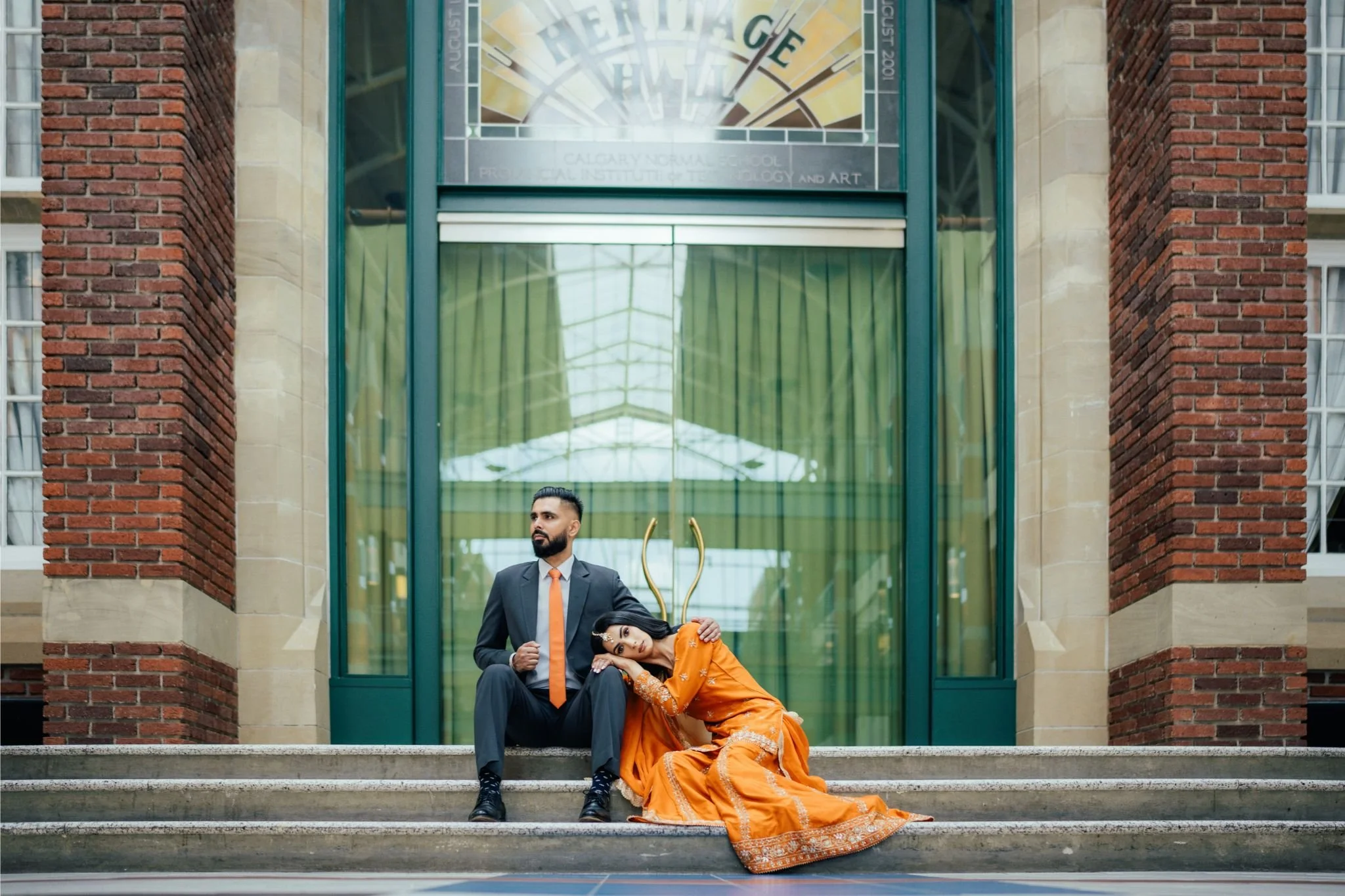 A man in a suit and an orange tie sitting on steps beside a woman in a traditional orange dress, who is resting her head on his shoulder and looking into the camera. They are outside a building with brick walls and large glass windows.
