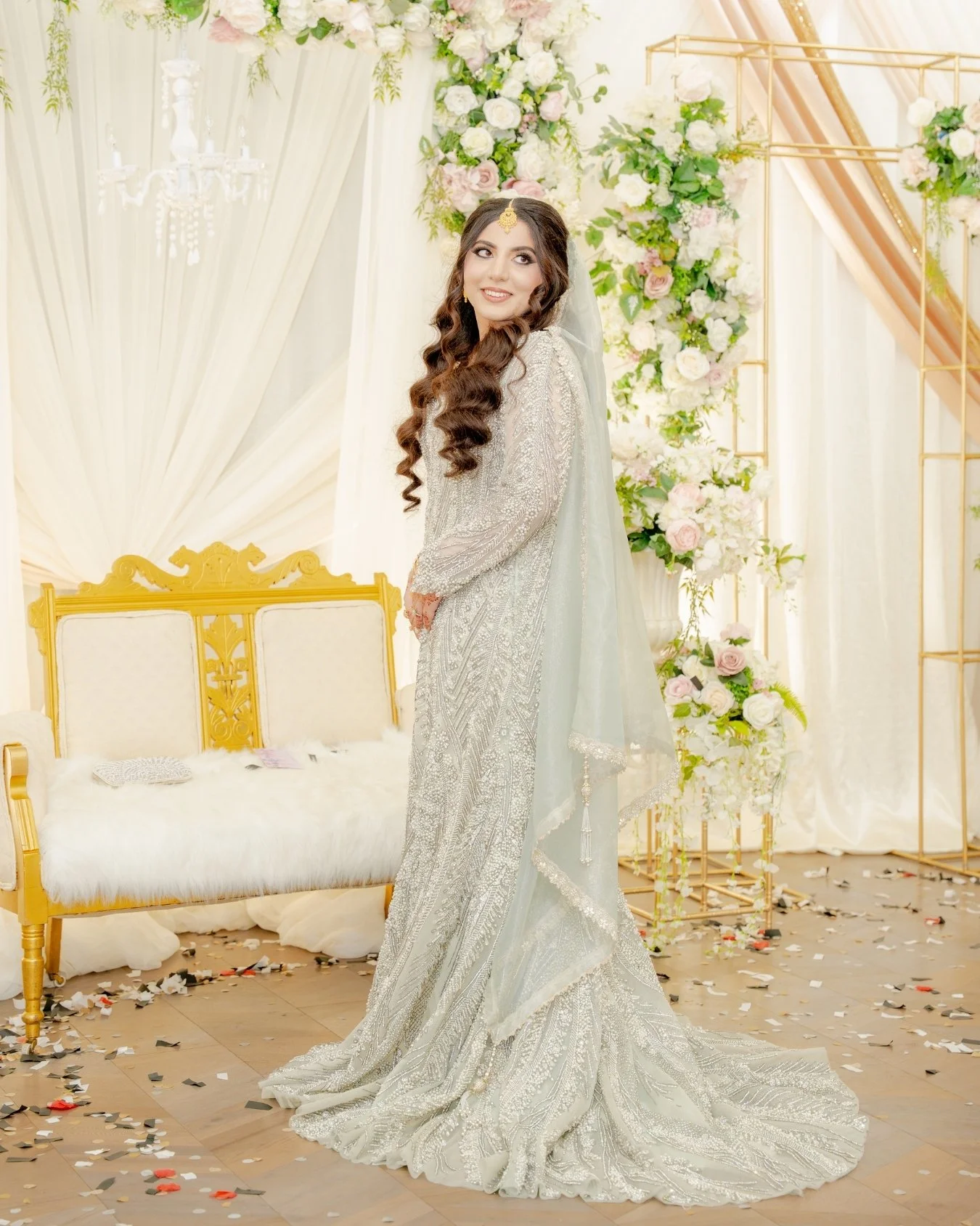 A bride in an ornate silver gown with intricate embroidery, standing in a decorated wedding venue with floral arrangements and gold accents. Nikkah bride
