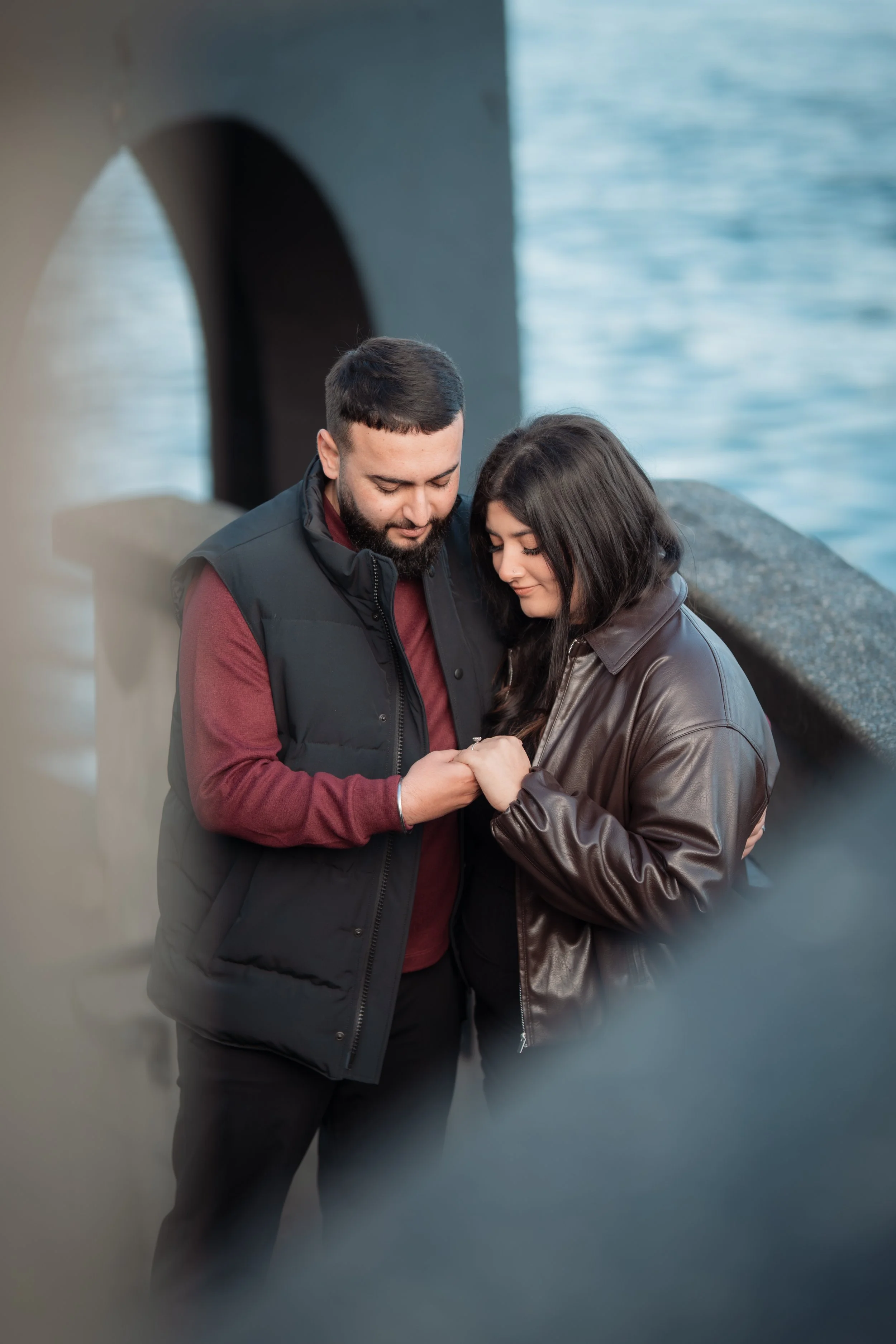 A young couple stands close together near water, holding hands and looking affectionately at each other.