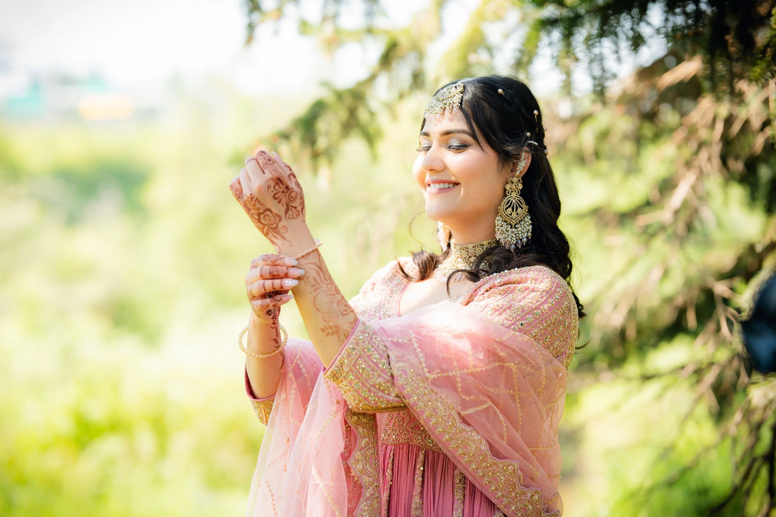 A woman dressed in traditional Indian attire, wearing jewelry and mehndi on her hands, smiling outdoors with green foliage in the background.