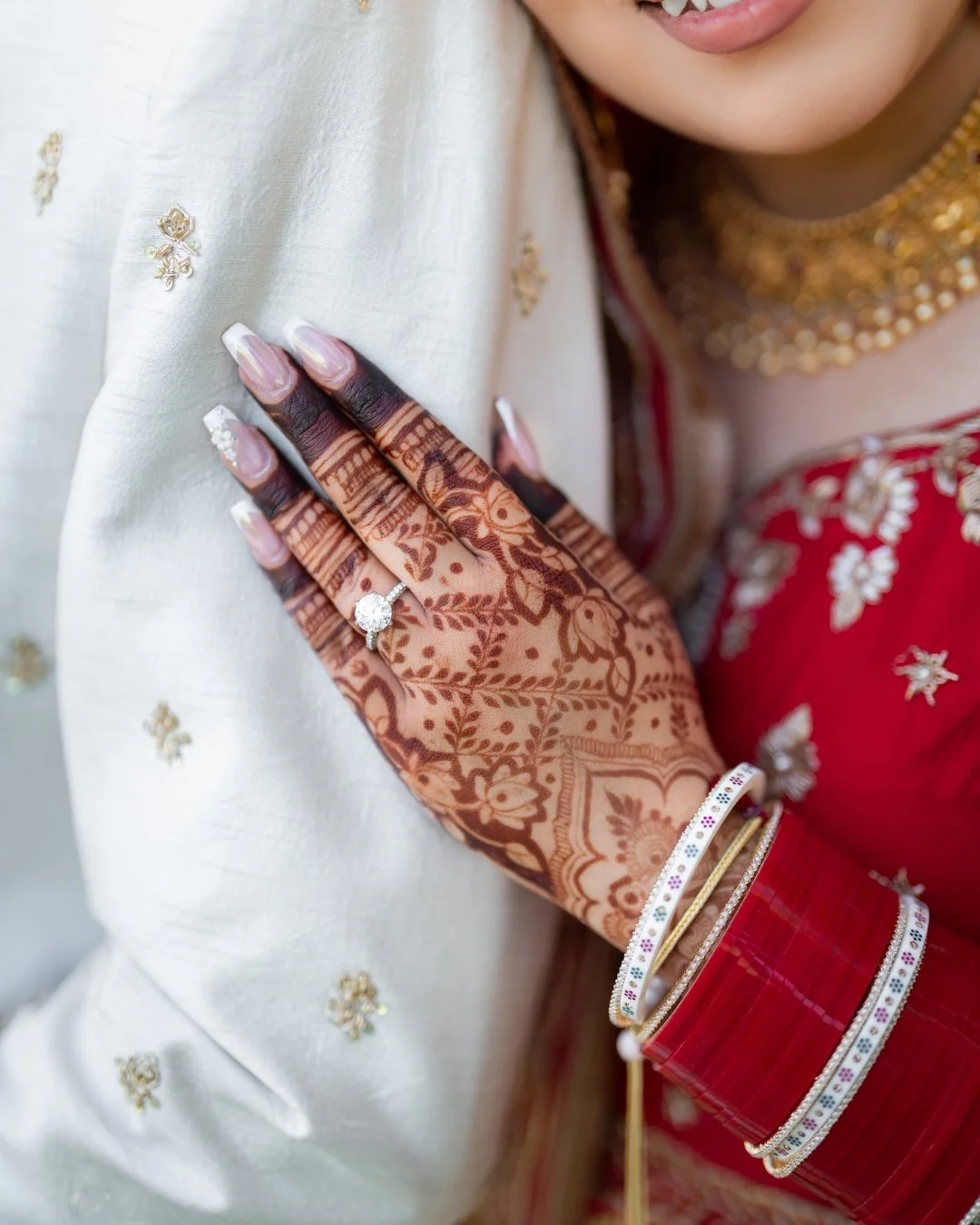 Close-up of a woman with henna-decorated hand wearing a diamond ring, resting on a person's shoulder dressed in white and red traditional Indian attire with gold jewelry and bangles.