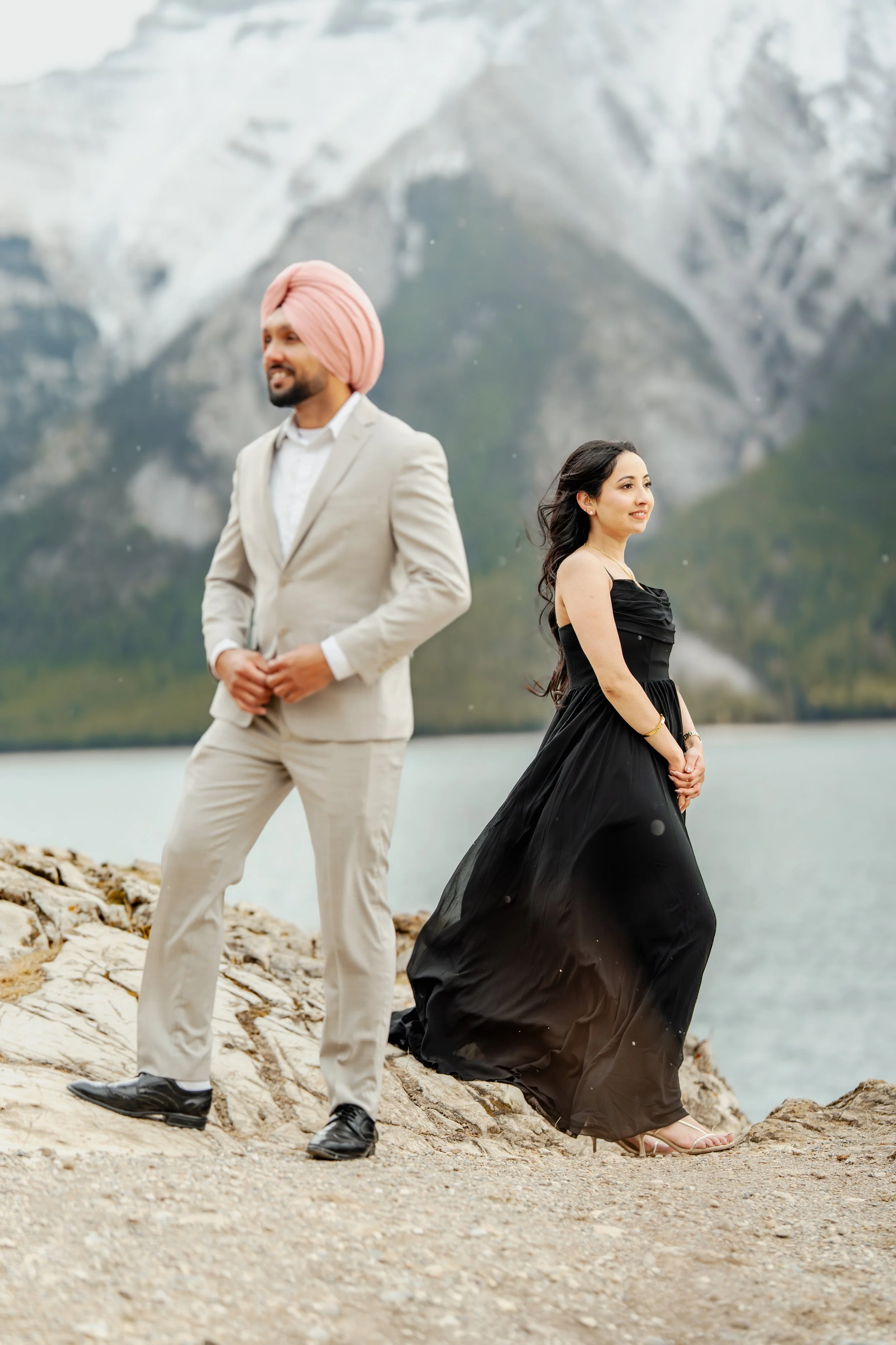 A man in a beige suit and pink turban stands on rocks near a lake, with a woman in a black dress in the background, overlooking mountains and water.