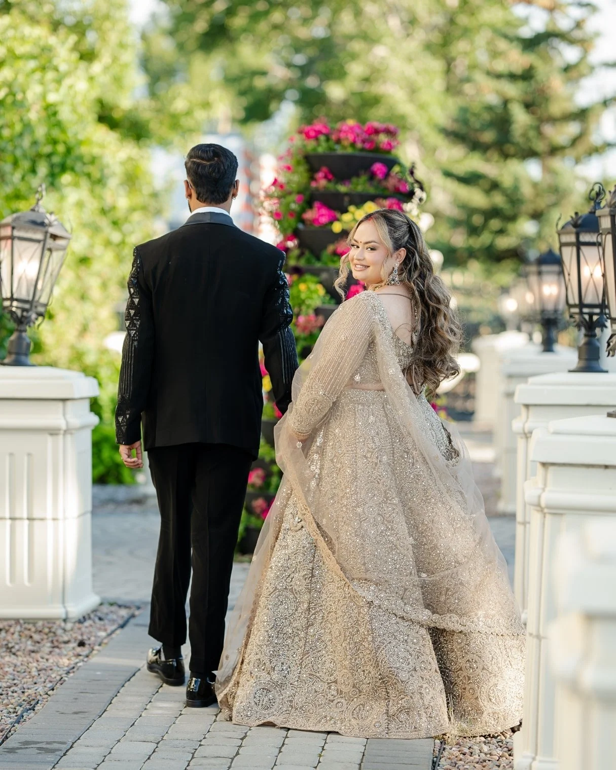 A woman in an ornate gown looks back and smiles at a man in a formal black suit walking ahead, set outdoors near decorative lamp posts and a colorful flower arrangement.