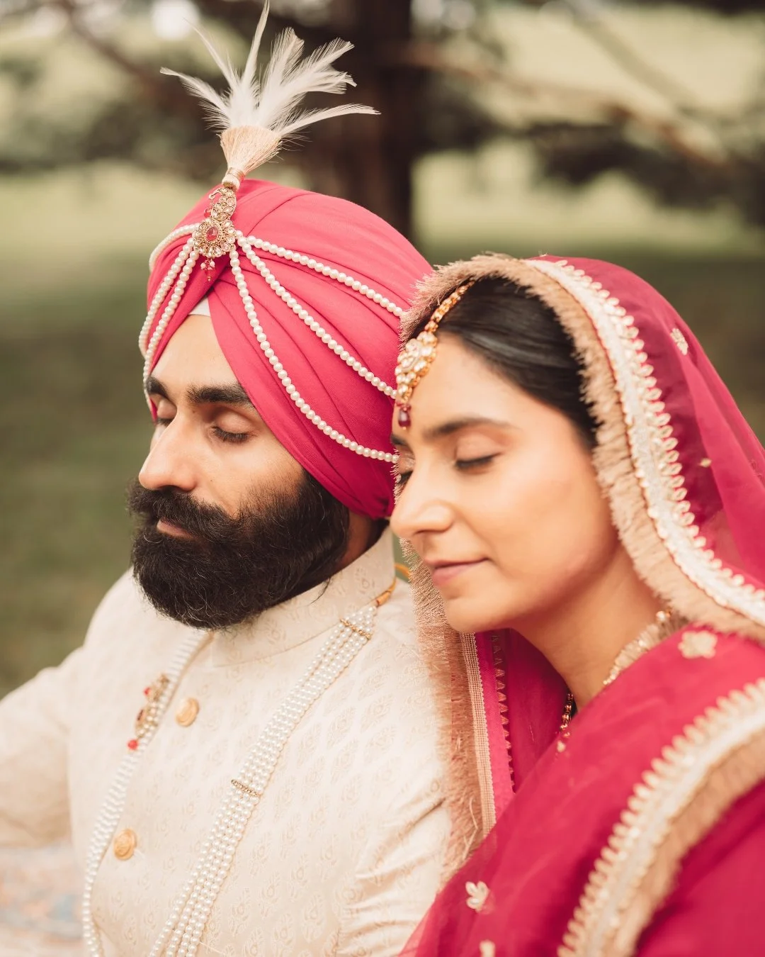 A man and woman dressed in traditional Indian wedding attire, sitting with their eyes closed, outdoors.