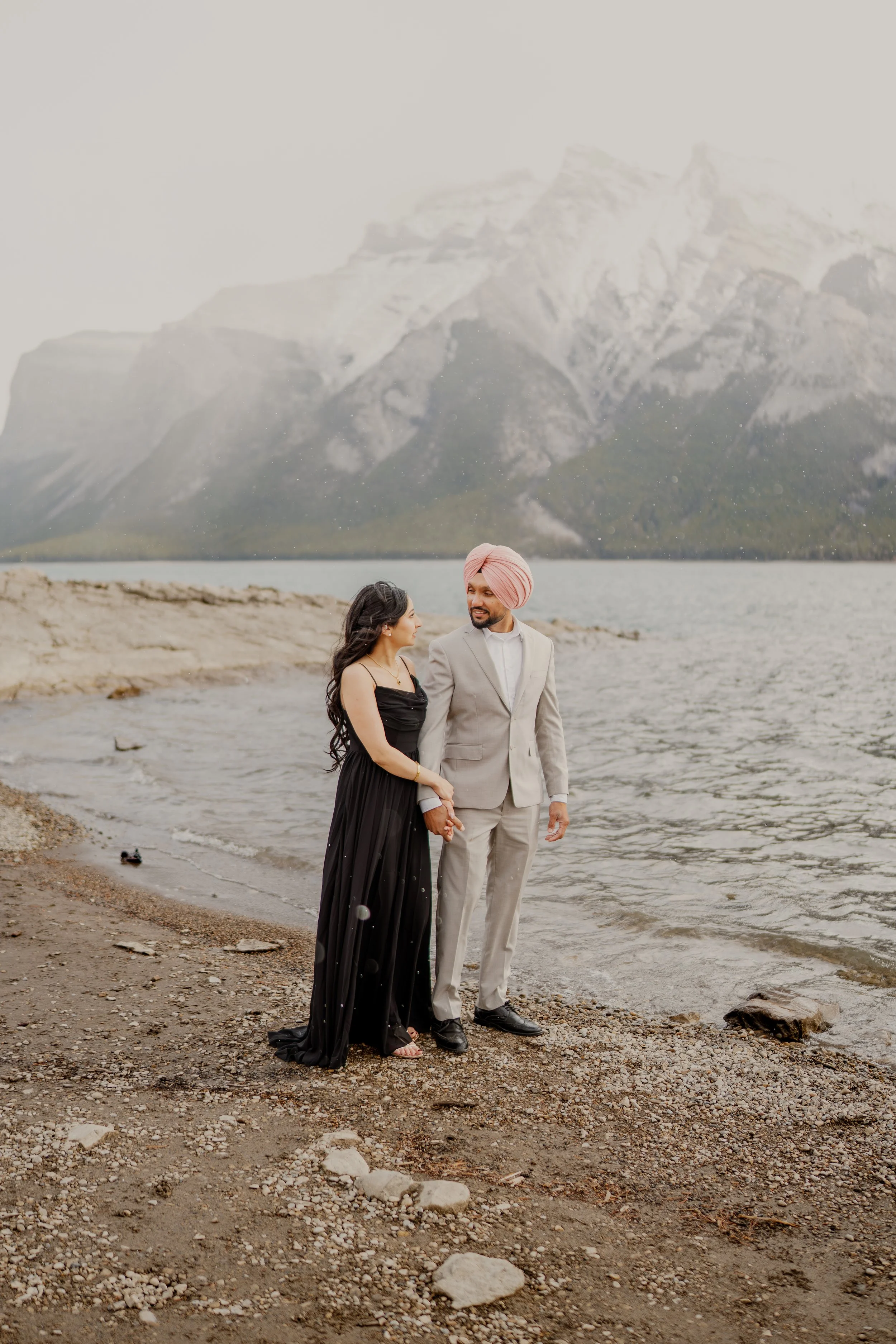 A couple holding hands on a rocky shoreline with mountains and a lake in the background, the woman is dressed in a black dress and the man in a light gray suit with a pink turban.