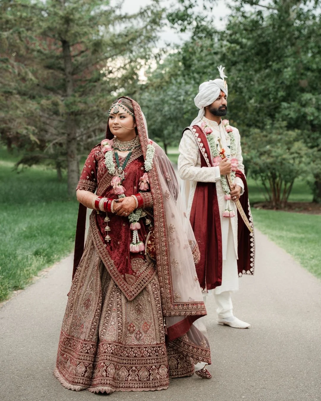 Indian bride and groom dressed in traditional wedding attire standing outdoors on a pathway, surrounded by greenery.