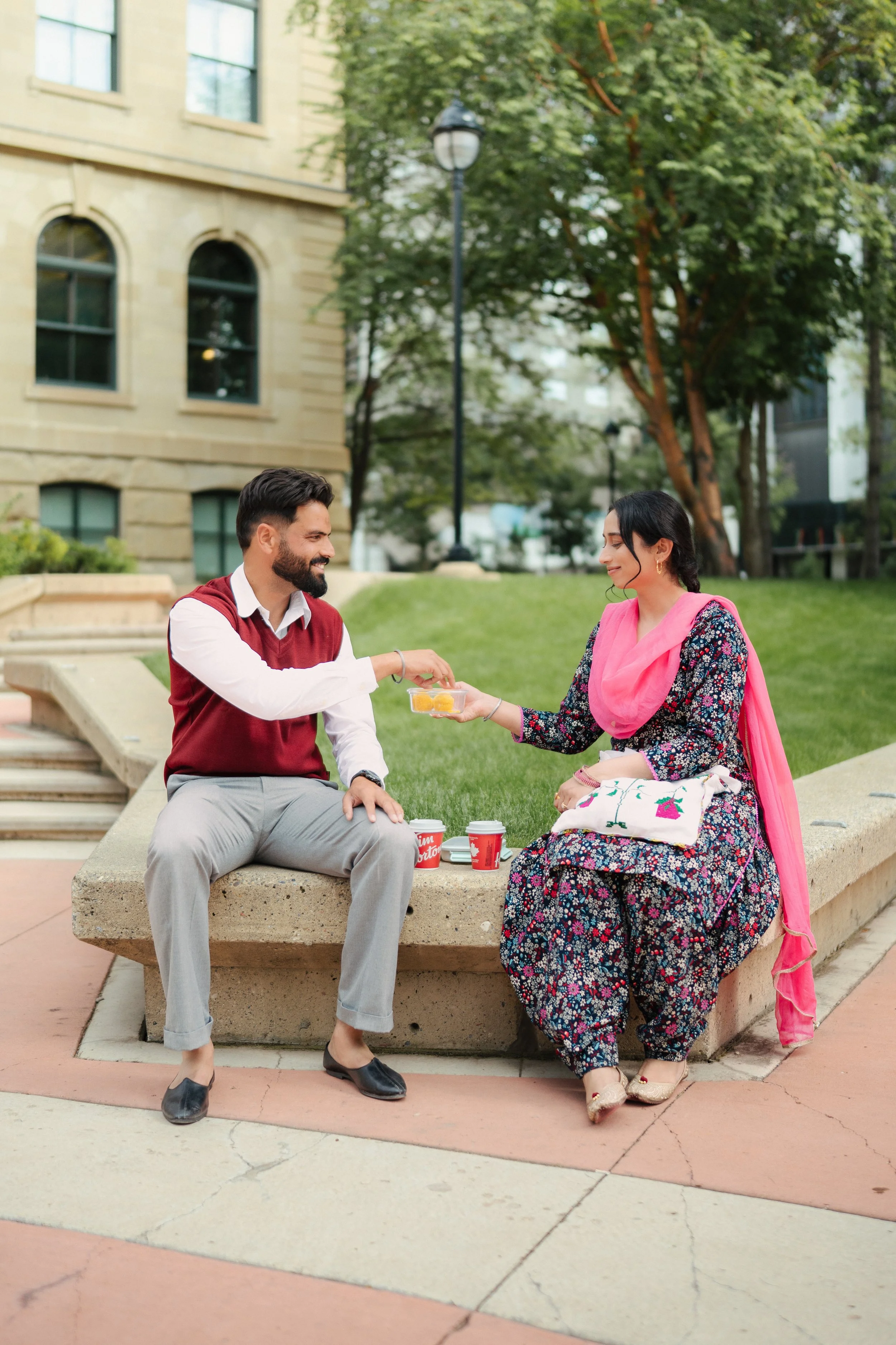 A man and a woman sitting on a concrete bench outdoors, exchanging food and drinks, with trees and a building in the background.
