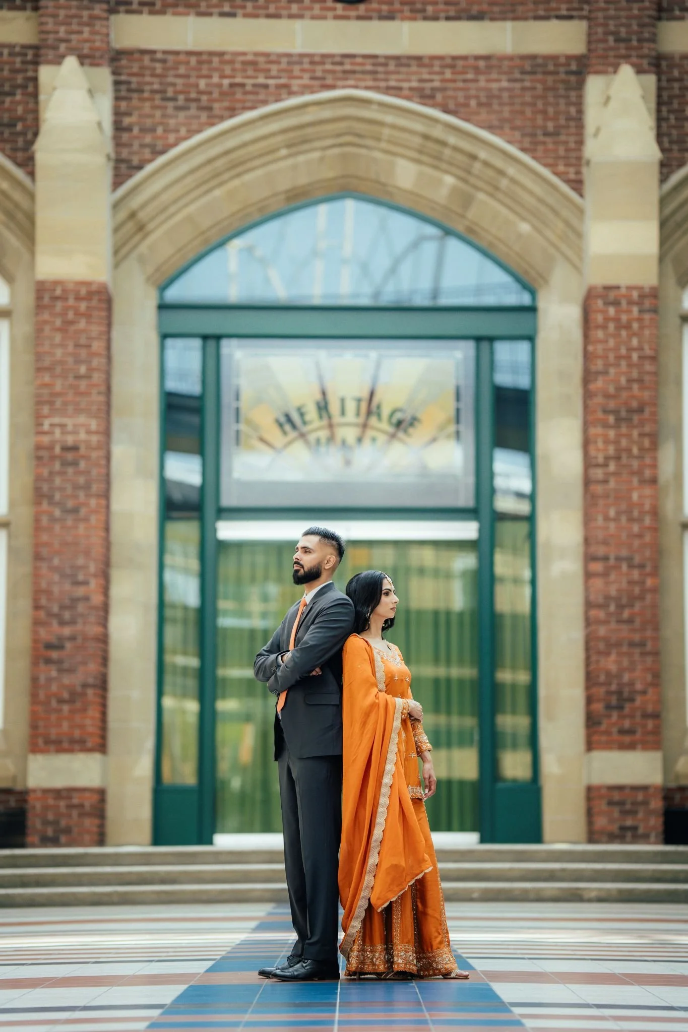 A man in a suit and a woman in an orange traditional dress are standing back to back with arms crossed outside a building with a sign that says 'Heritage.'