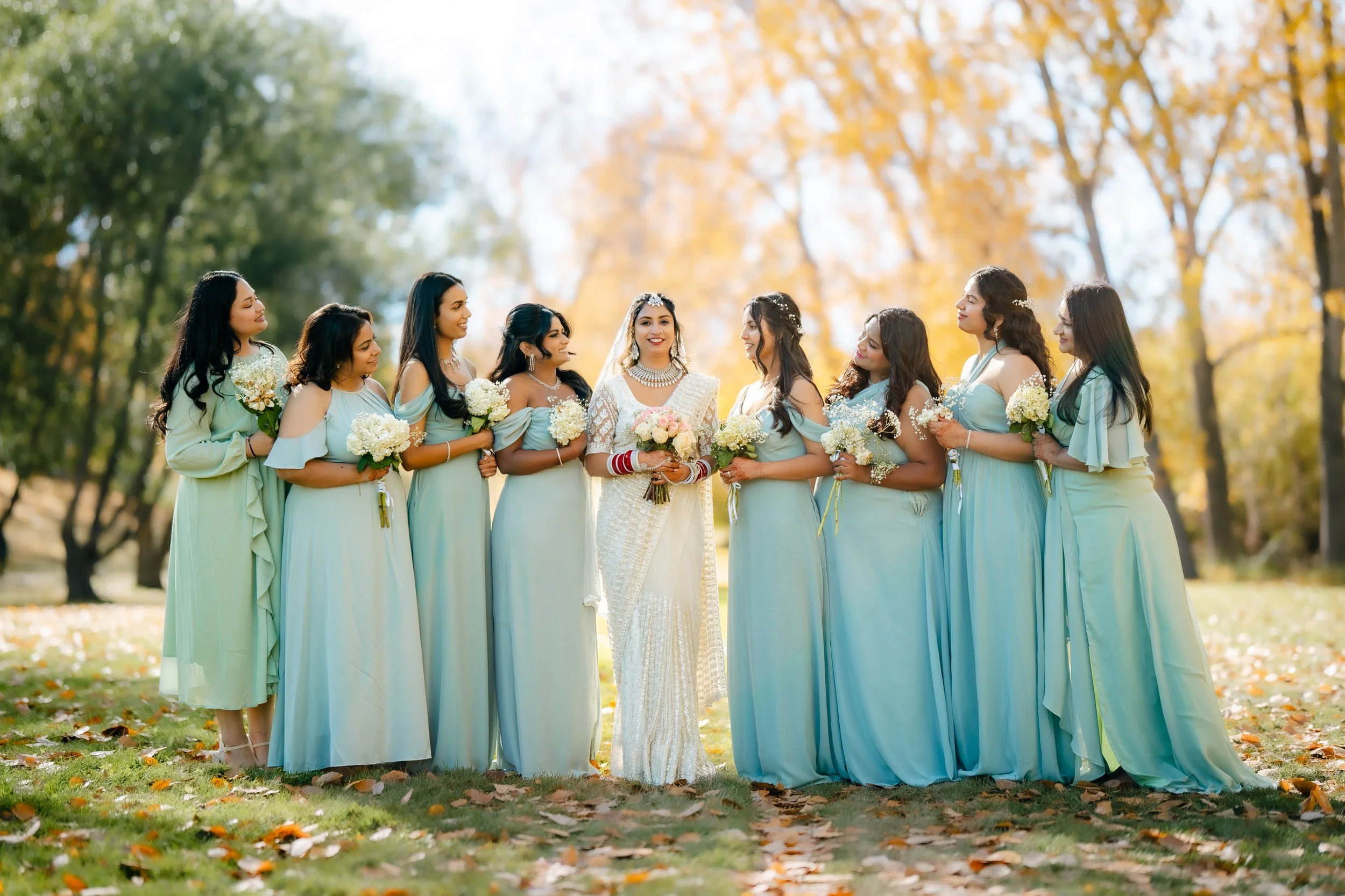 A bride in traditional wedding attire surrounded by bridesmaids in light blue dresses, standing outdoors in a park with autumn foliage and fallen leaves.