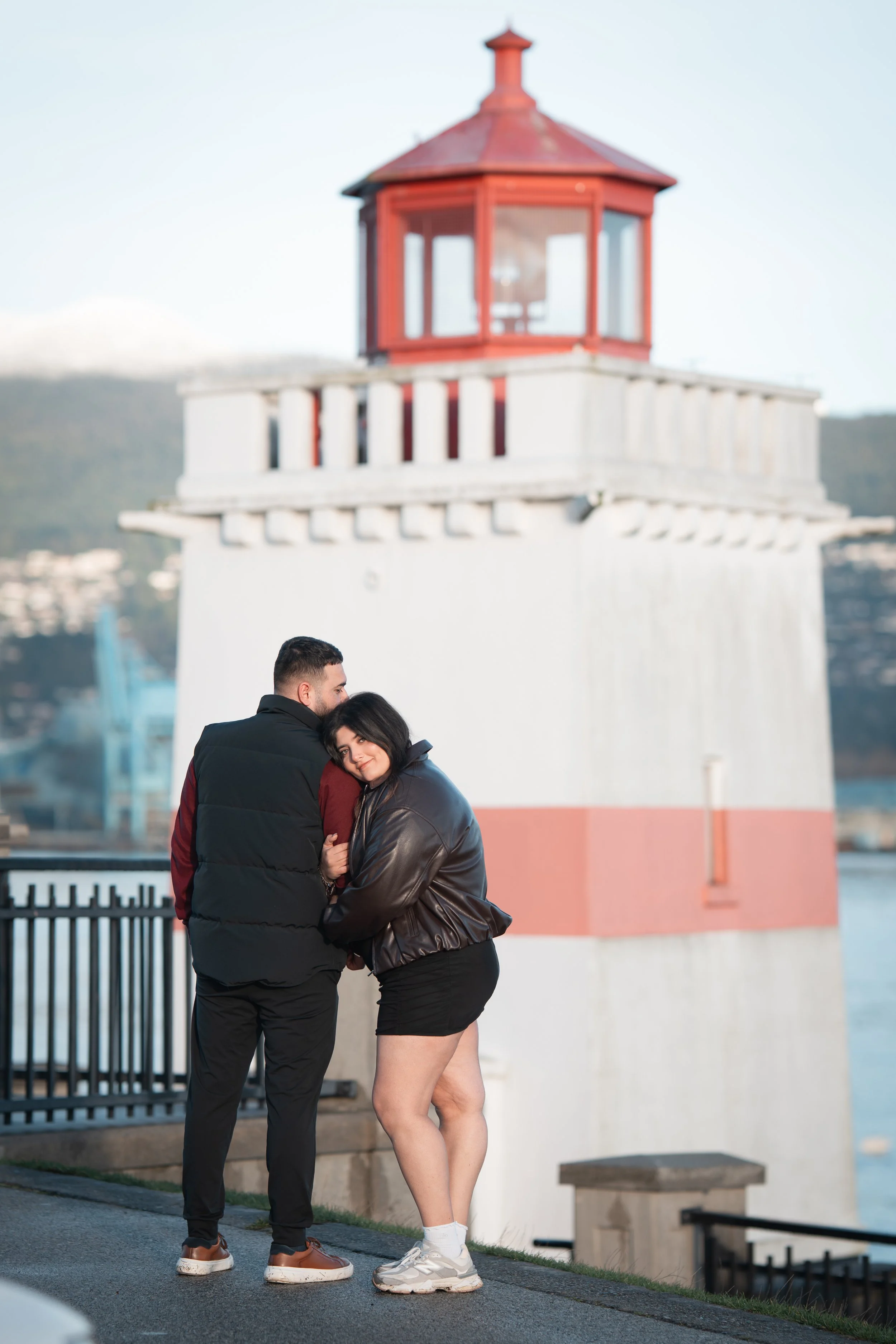 A couple hugging near a lighthouse during daytime, with the man facing away and the woman leaning into him, smiling.