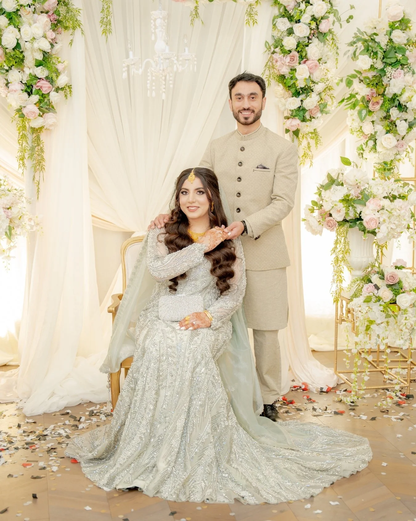 A newlywed couple at their wedding reception, with the bride seated in an ornate silver wedding dress and the groom standing behind her, both smiling. The background features elegant white and pink floral arrangements, draped white curtains, and a ch
