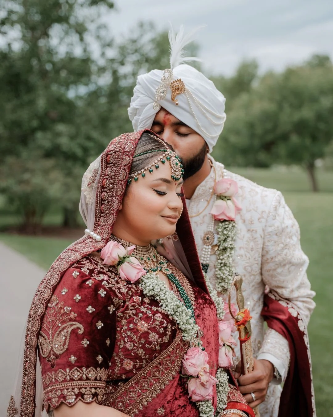 A couple dressed in traditional Indian wedding attire, the groom kissing the bride on the forehead outdoors with trees in the background.