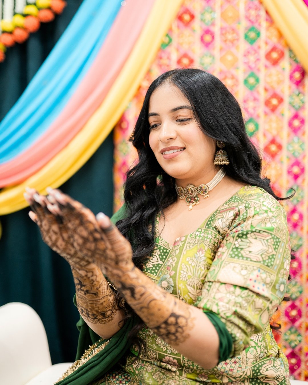 A woman in traditional Indian attire, with henna decorations on her hands, smiling while looking down at her hands against a colorful, decorated background. Bridal Mehndi 
