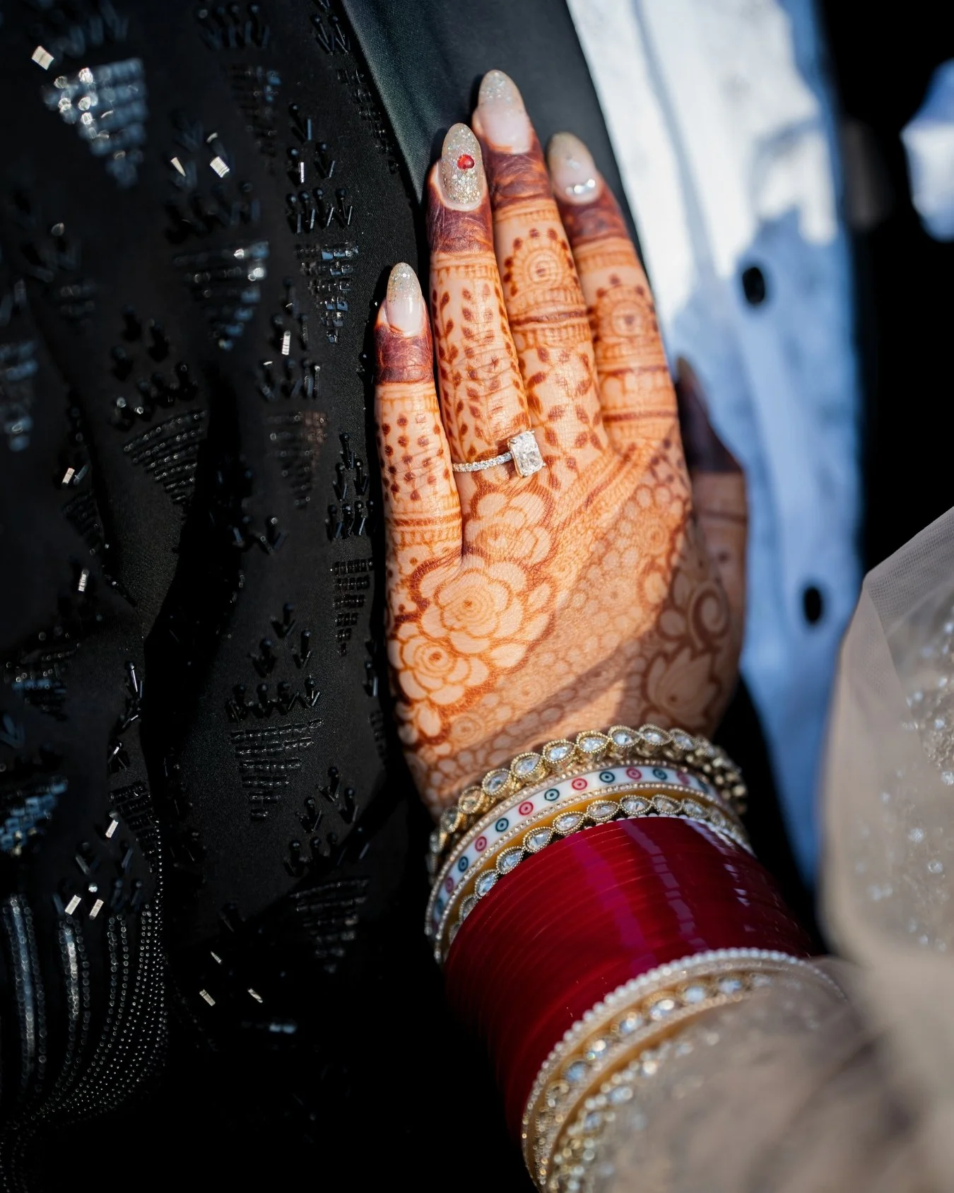 Close-up of a hand with mehndi and rings resting on a decorative black surface, surrounded by colorful bangles.