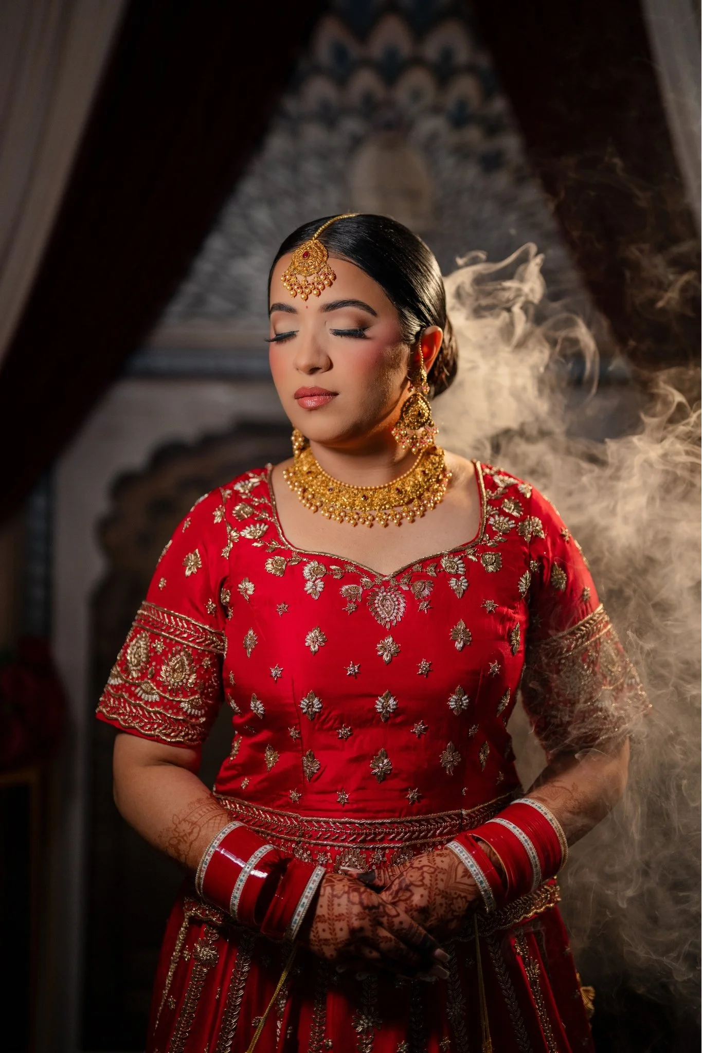 A woman in traditional Indian bridal attire wearing a red dress with gold embroidery, gold jewelry, and henna on her hands, standing with her eyes closed.