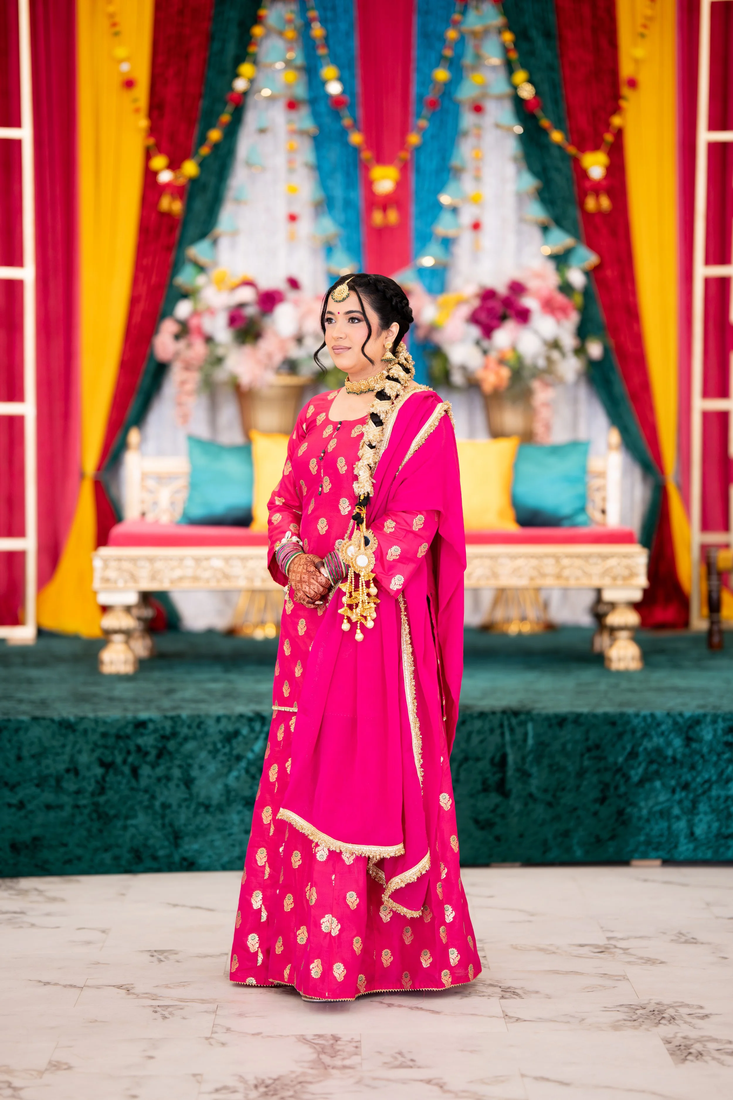 A woman dressed in traditional Indian pink attire, standing in front of a colorful decorated background with curtains, flowers, and cushions, likely at a celebration or wedding. Bride side jaggo
