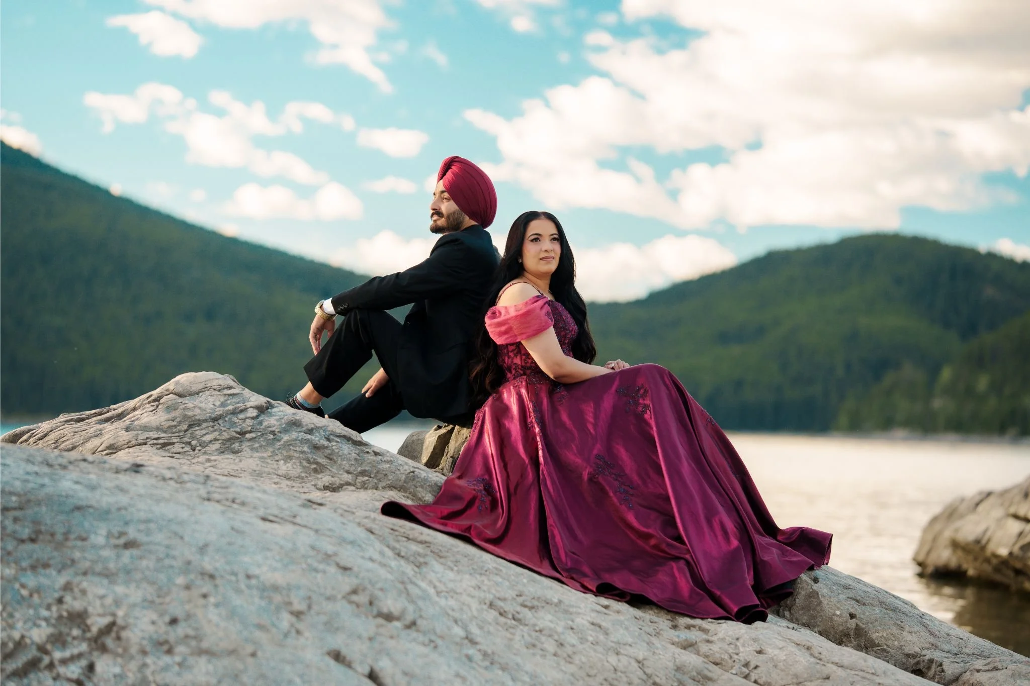 A man wearing a black suit with a red turban and a woman in a purple gown sitting on rocks near a body of water with mountains and a cloudy sky in the background.