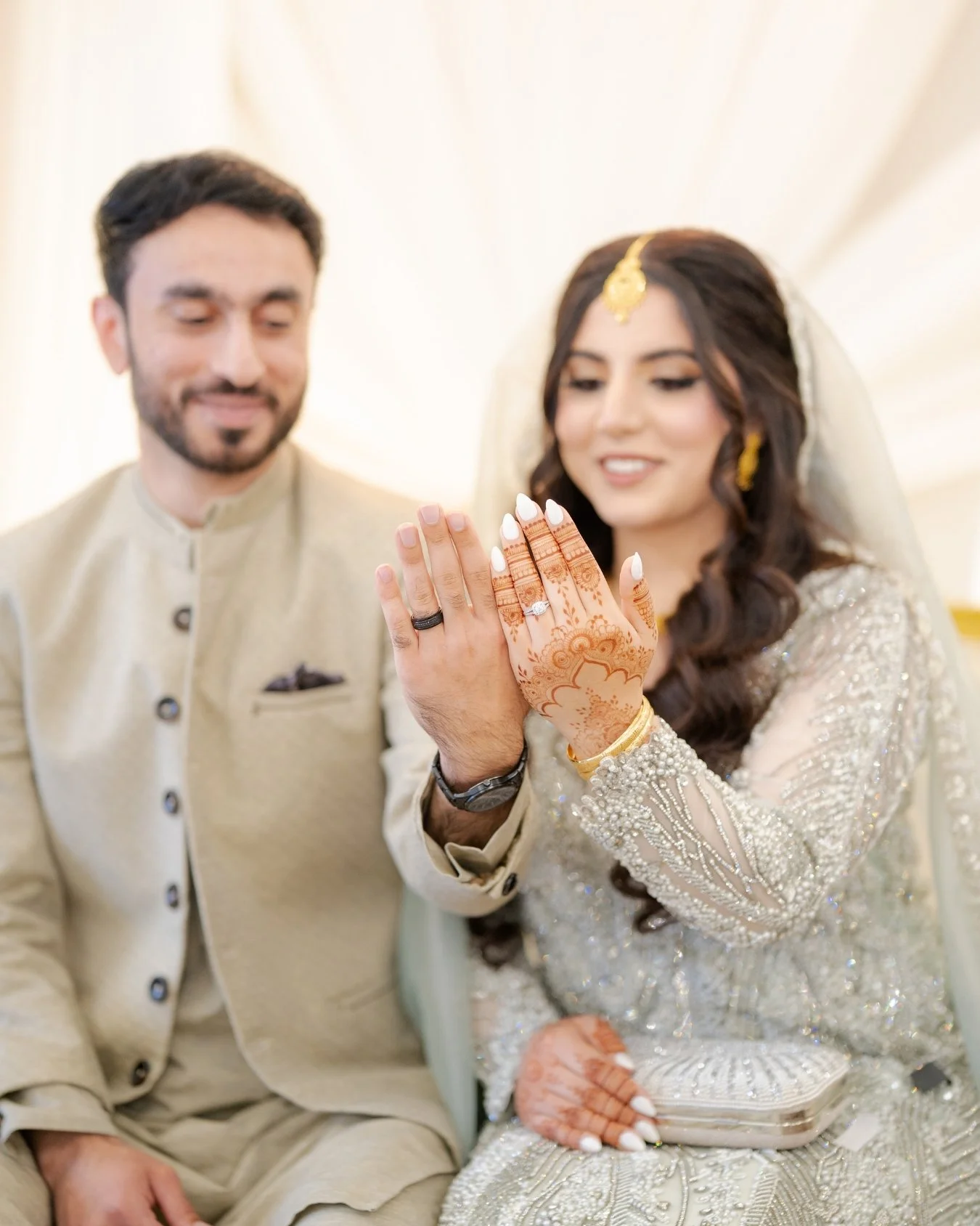 A couple celebrating their wedding, with the bride displaying her wedding ring and henna decorated hands, wearing traditional attire, and the groom beside her, smiling.