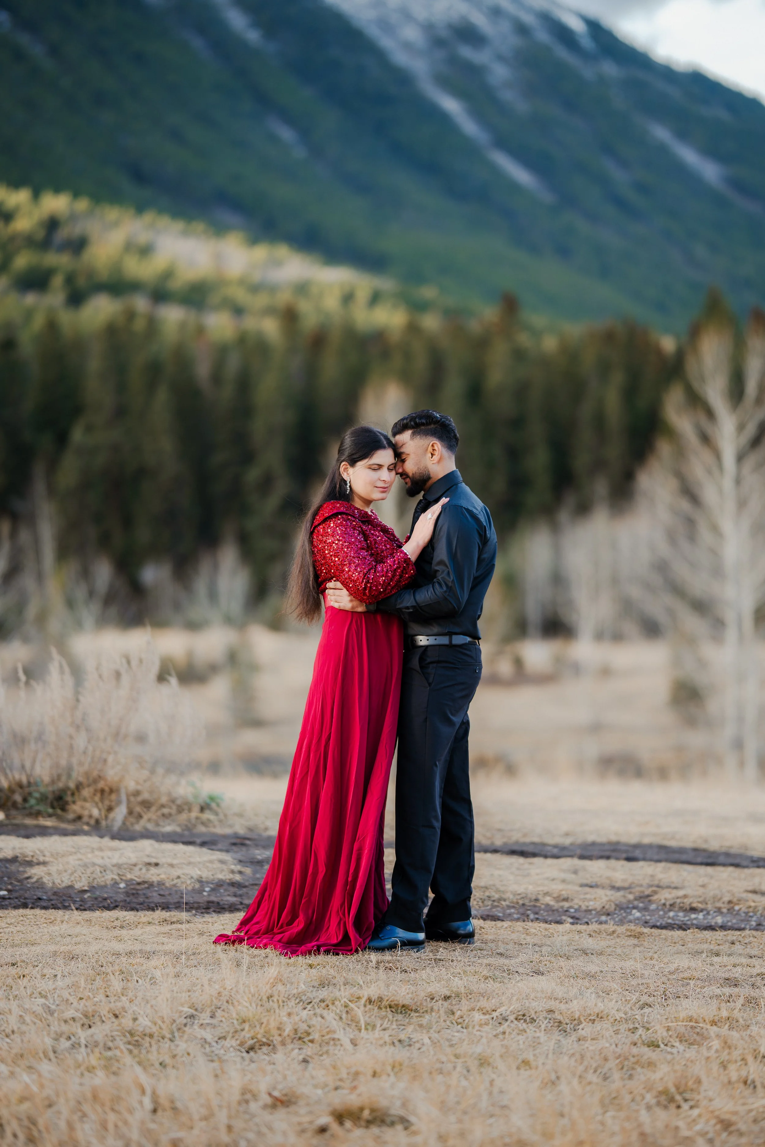 A couple embracing outdoors, the woman in a long red dress and the man in a black shirt, with mountains and trees in the background.
