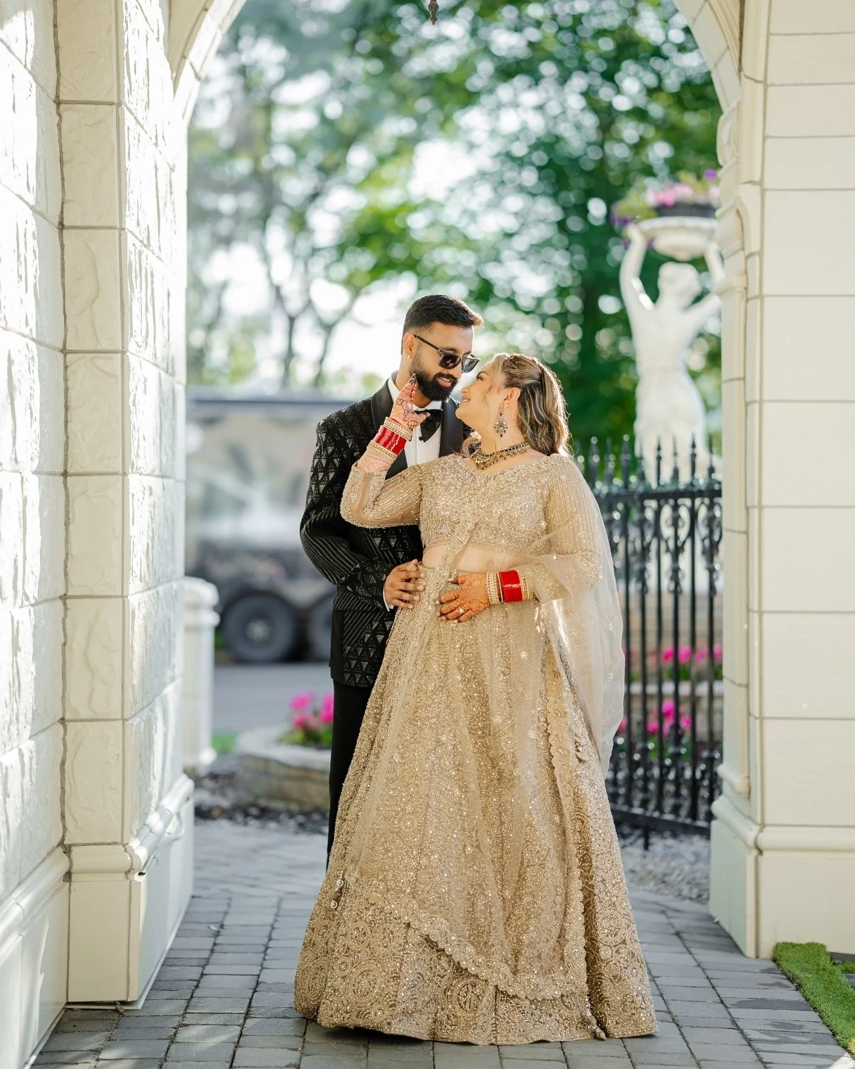 A couple dressed in traditional Indian attire, standing close together under an archway, with trees and a statue in the background.