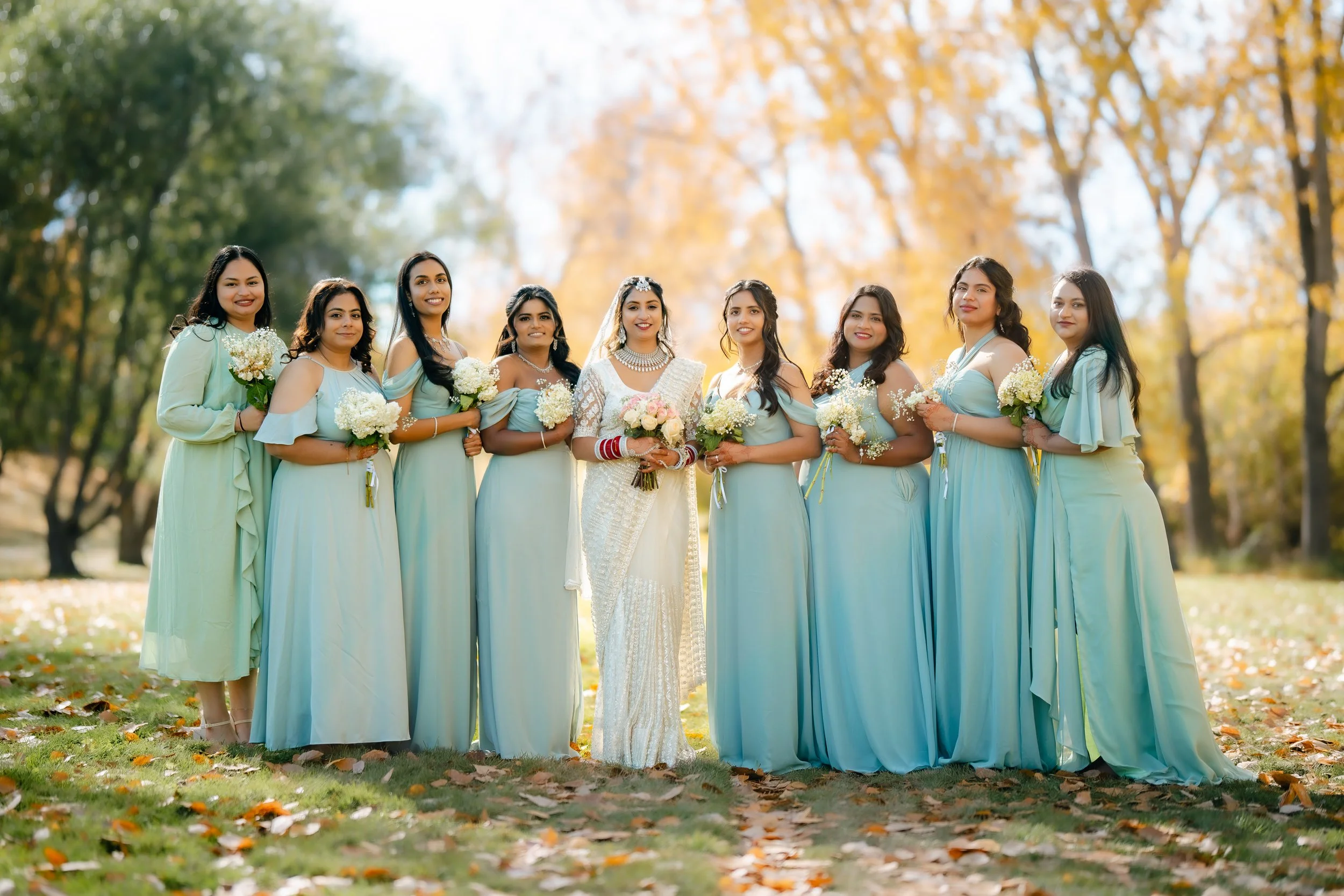 A bride and nine bridesmaids in matching light blue dresses holding bouquets in a park with autumn trees and fallen leaves. Western wedding