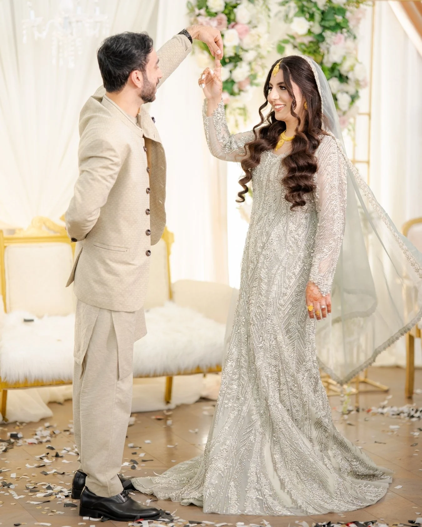 A bride and groom at their wedding ceremony. The groom is holding the bride's hand, and they are smiling. The bride is wearing a detailed, silver wedding dress with a veil, and the groom is dressed in a cream-colored suit. There are flower arrangemen