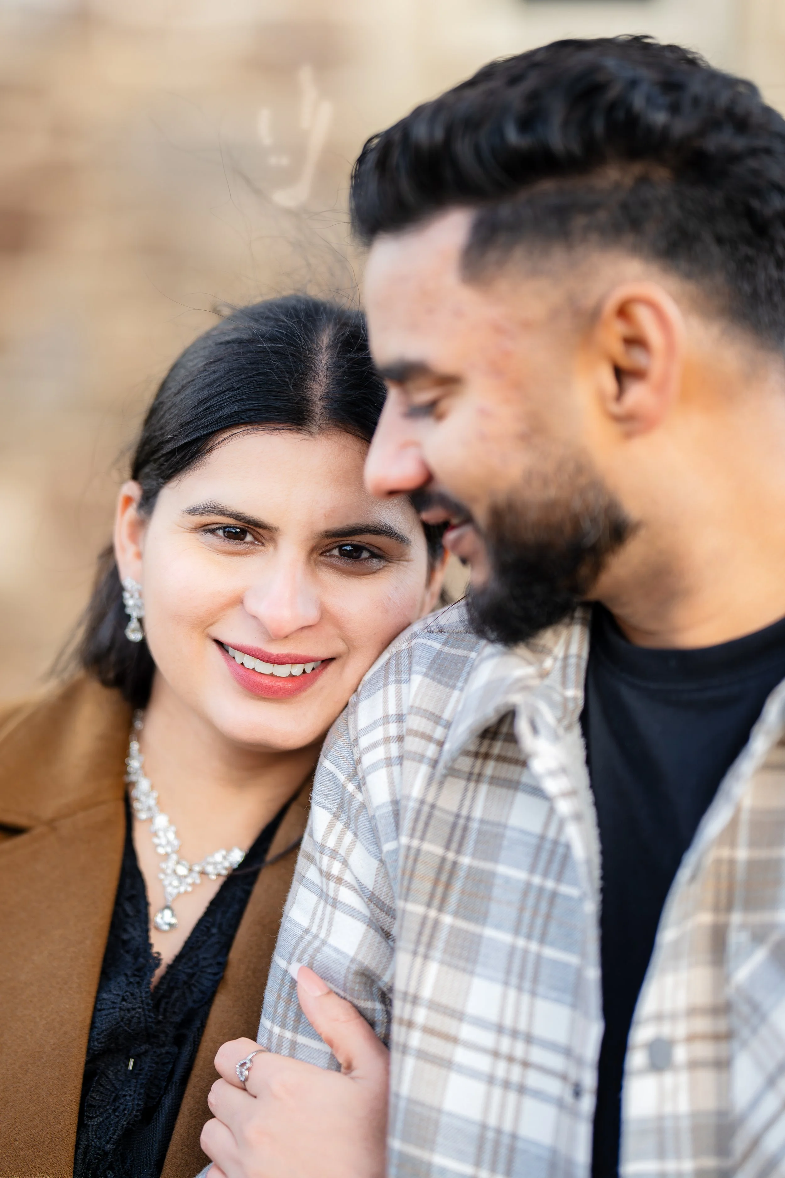 Smiling woman with dark hair and jewelry leaning against a bearded man with dark hair and a plaid shirt, outdoors.