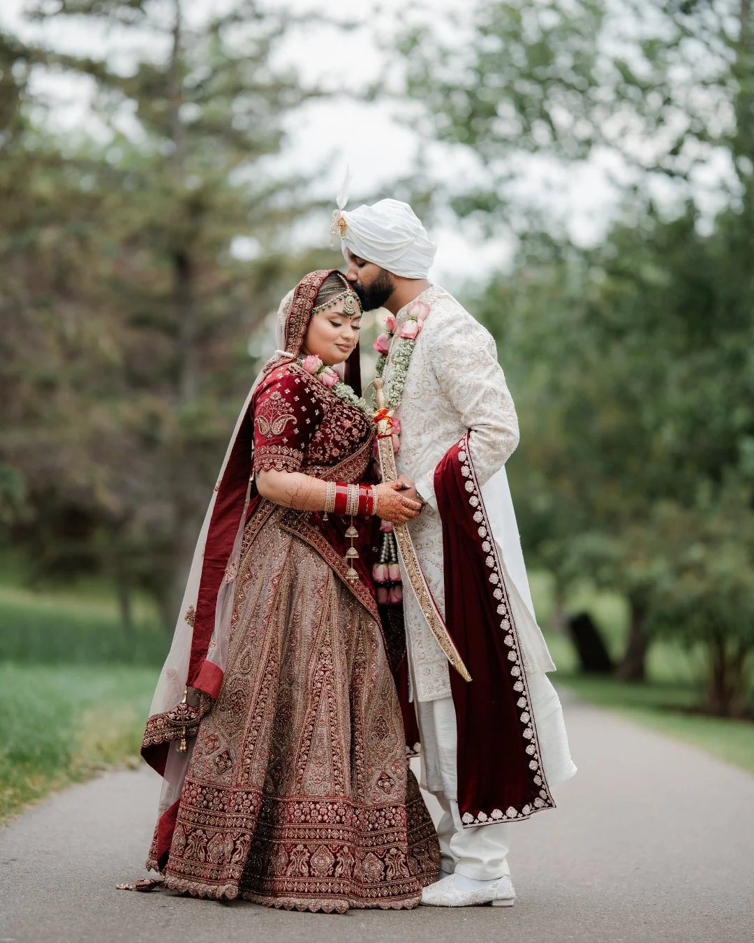 An Indian couple dressed in traditional wedding attire standing outdoors on a pathway surrounded by trees, sharing an intimate moment.