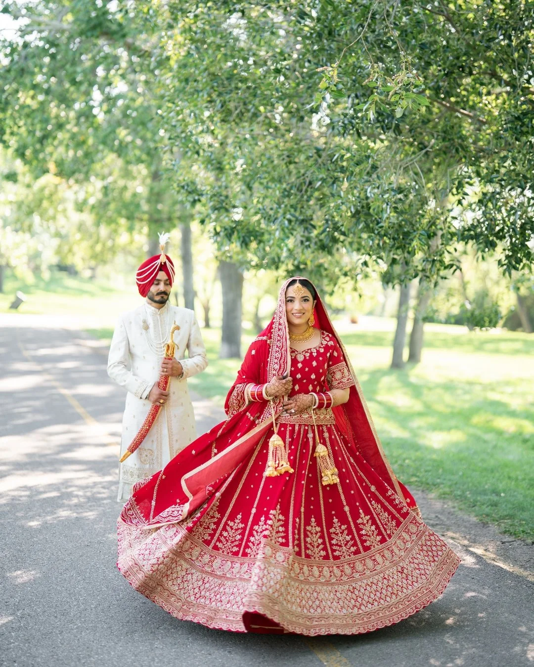Indian bride in red and gold wedding dress holding a dupatta, walking on a road with groom in white traditional attire and red turban in a park.
