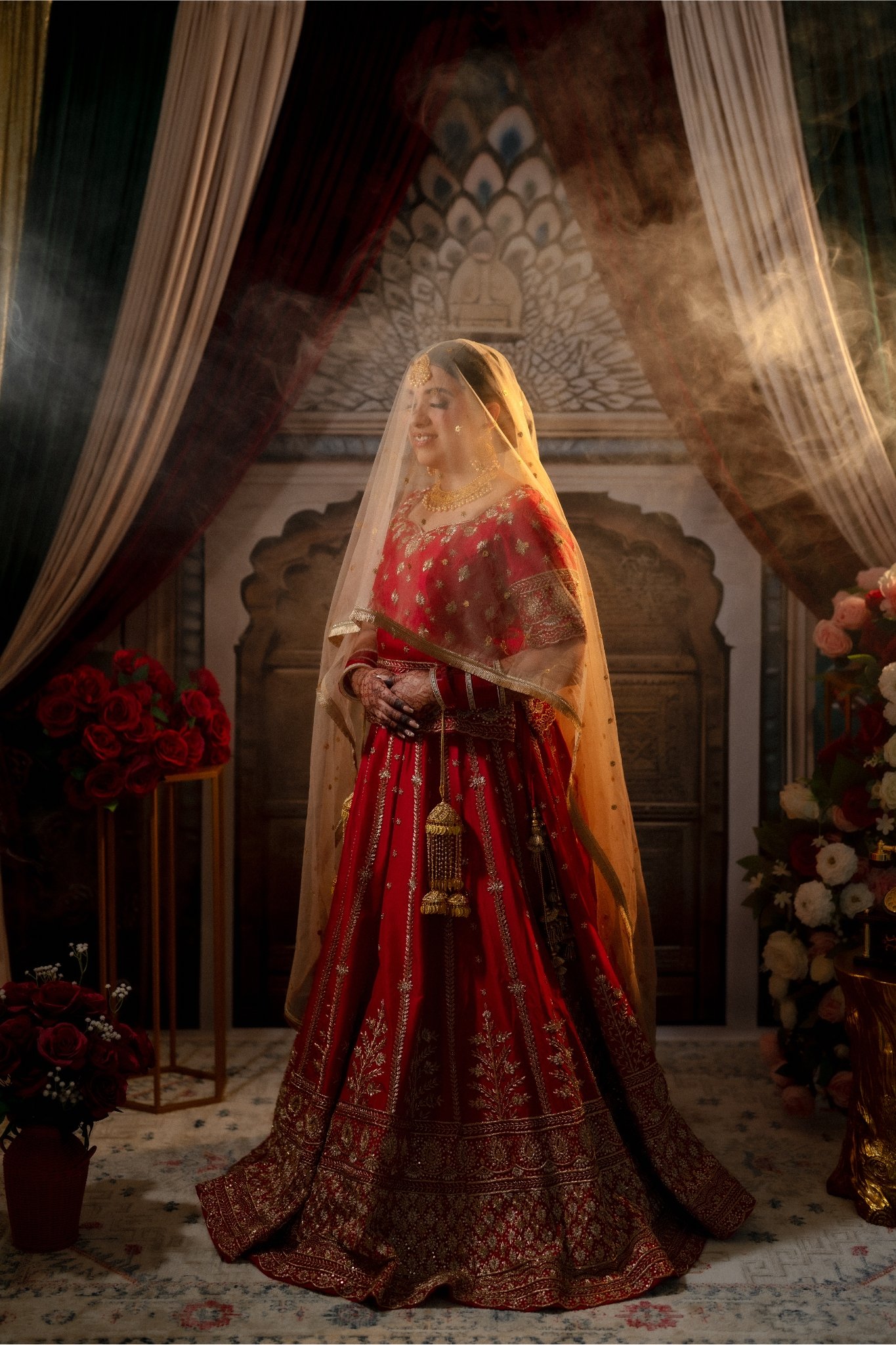 A bride in traditional Indian red bridal attire with jewelry, standing in a decorated room, surrounded by flowers.