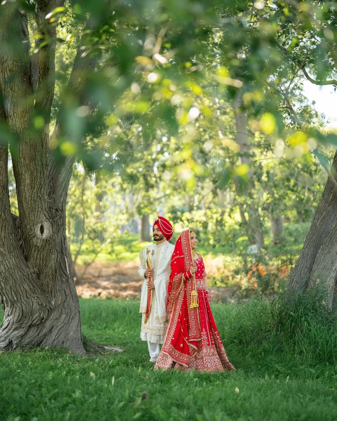 A couple dressed in traditional Indian wedding attire standing outdoors near large trees and green grass, with the woman in a red bridal outfit and the man in a white outfit with a red turban.