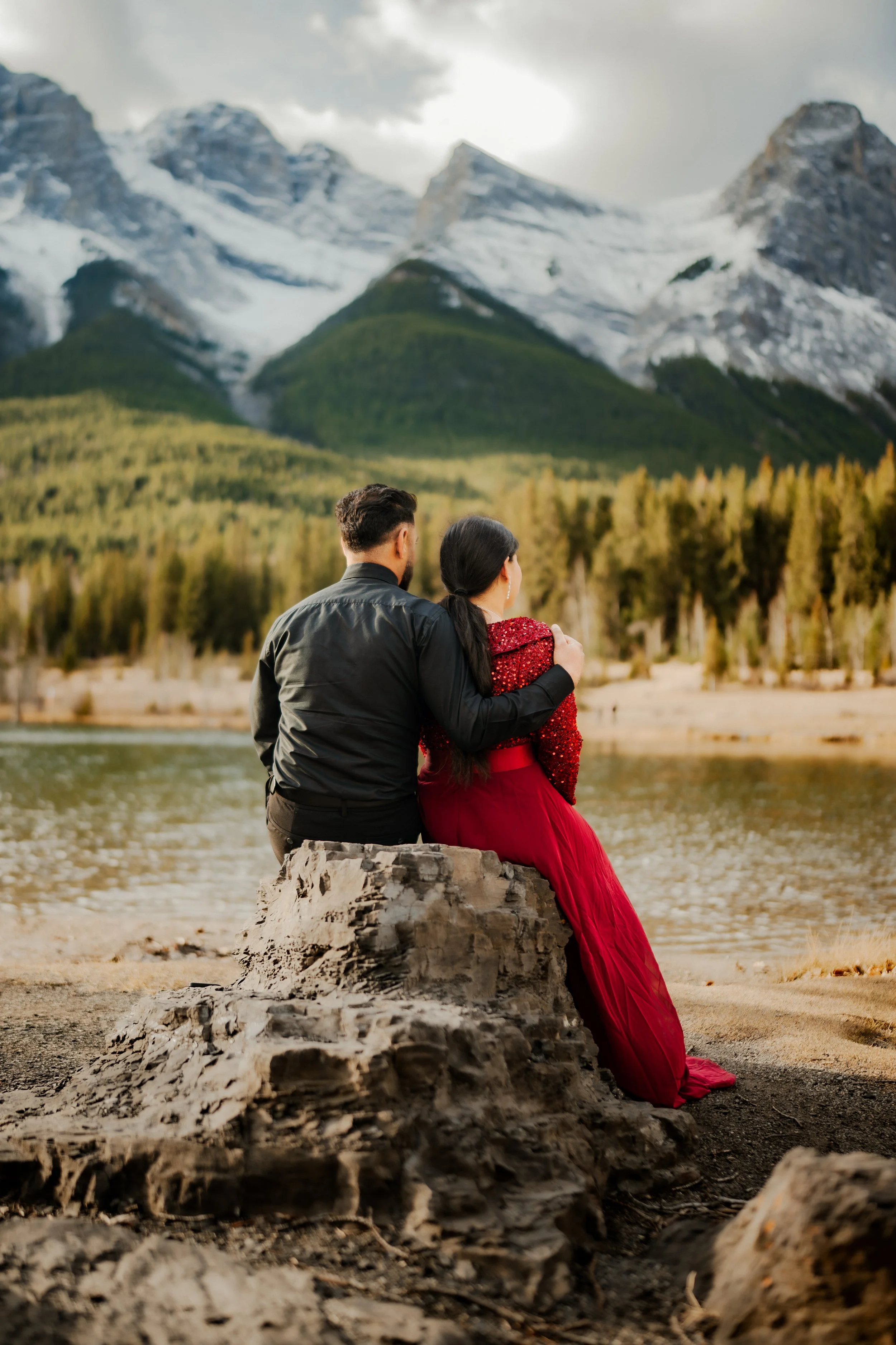 A couple sitting on a rock by a lake, surrounded by trees and mountains, watching the scenery.