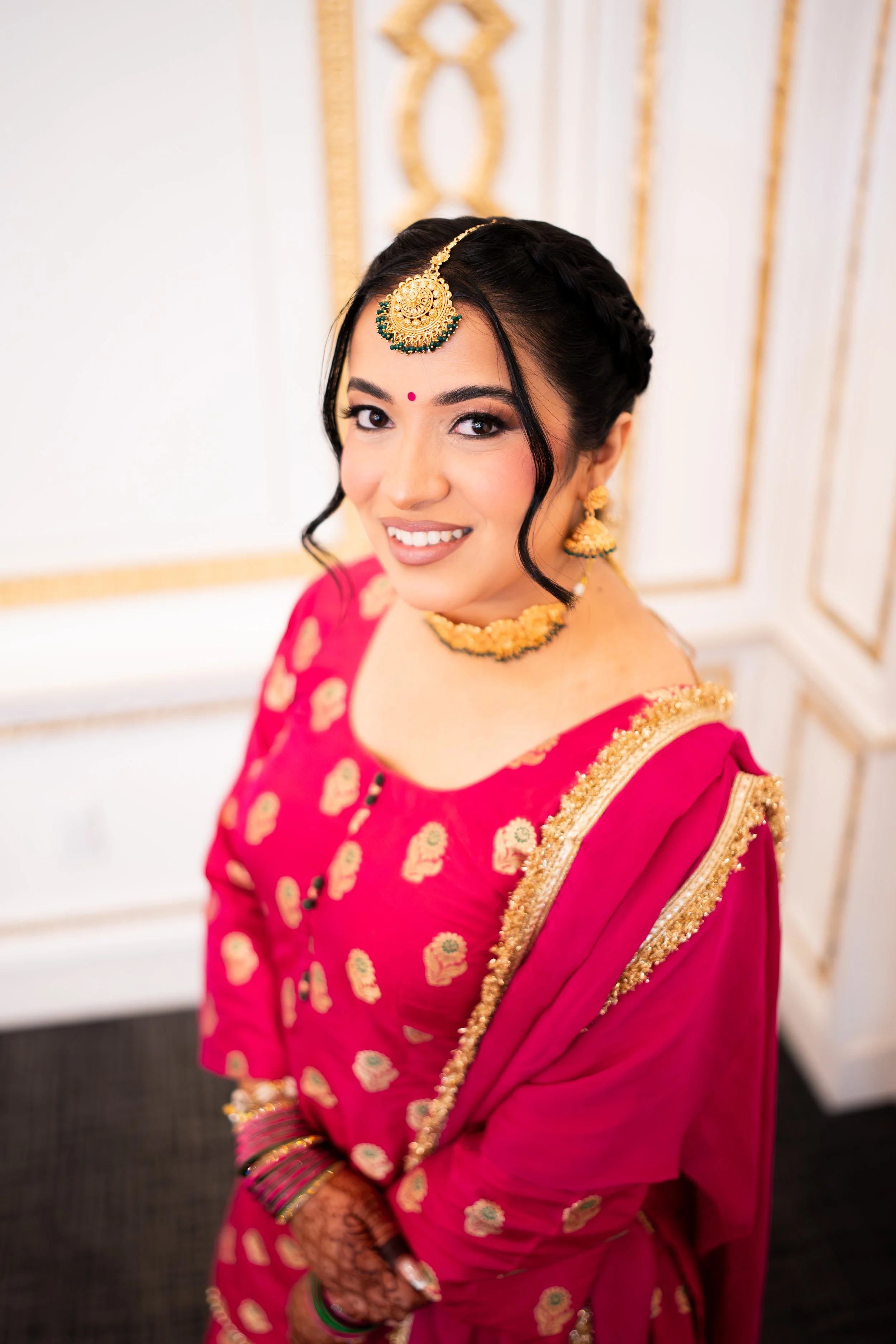 A woman dressed in traditional Indian attire, wearing a pink saree with gold embroidery, gold jewelry, and henna on her hands, smiling at the camera.