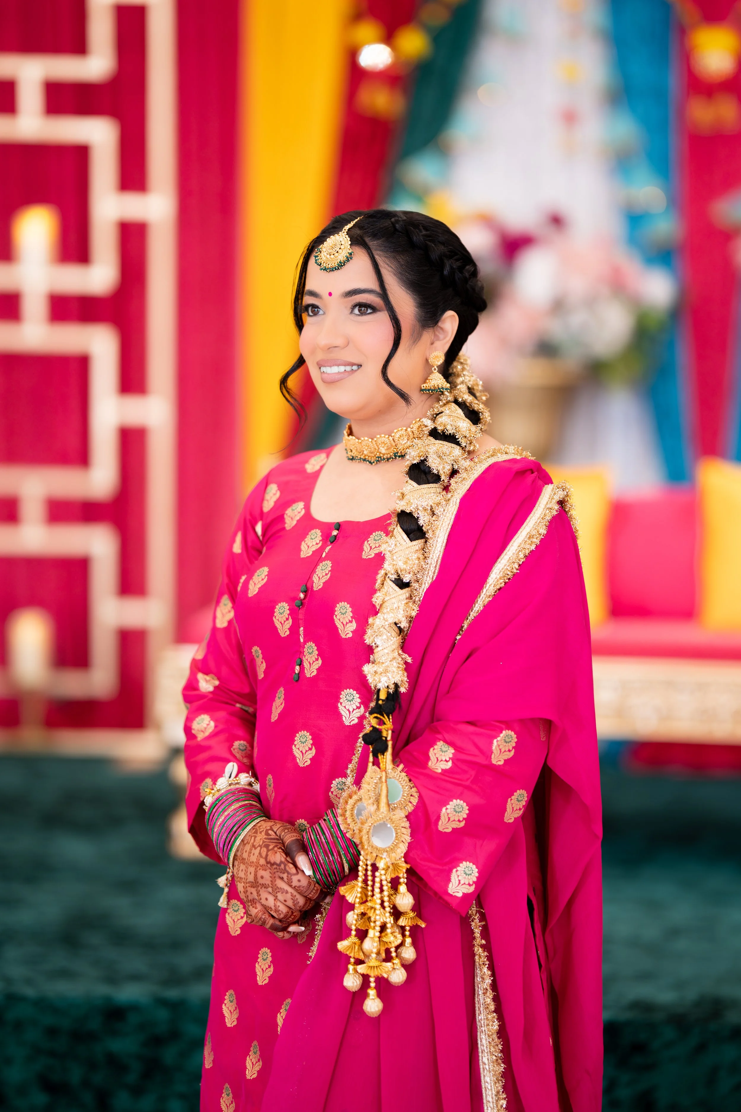 A woman dressed in traditional Indian attire, wearing a vibrant pink saree embellished with gold embroidery, accessorized with jewelry including earrings, necklace, and bangles, standing in a decorated setting with colorful drapes and flowers.