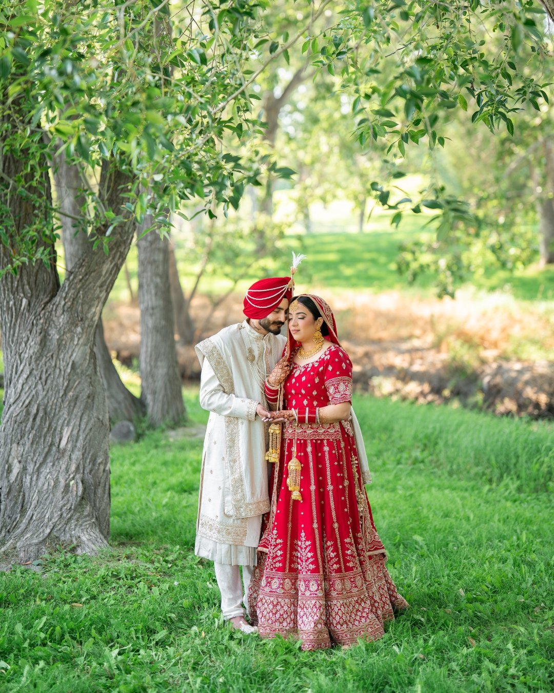 A traditional Indian wedding photo showing a bride and groom in colorful attire standing under a large leafy tree in a lush green outdoor setting.