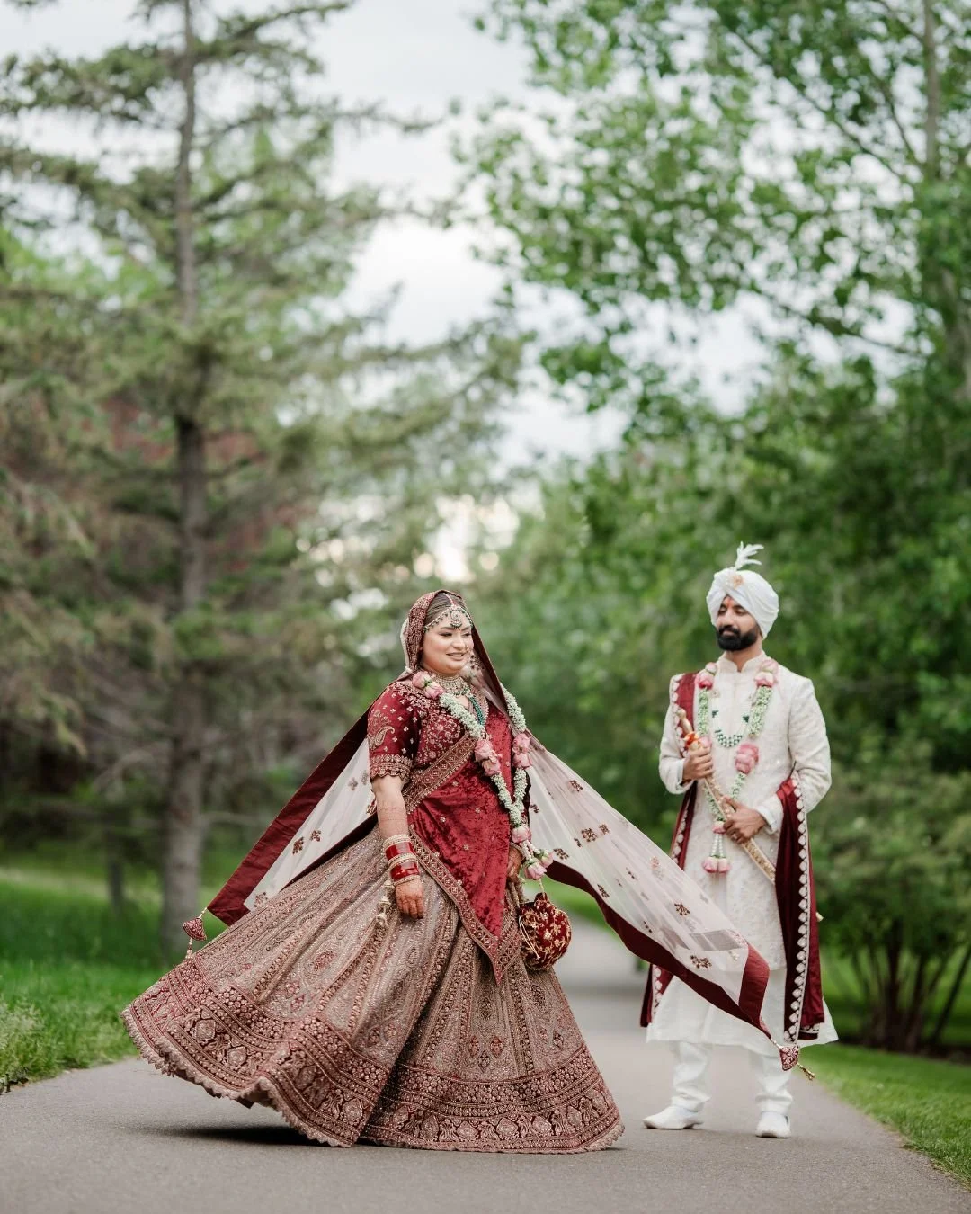 Indian bride and groom in traditional wedding attire standing outdoors on a pathway surrounded by green trees.