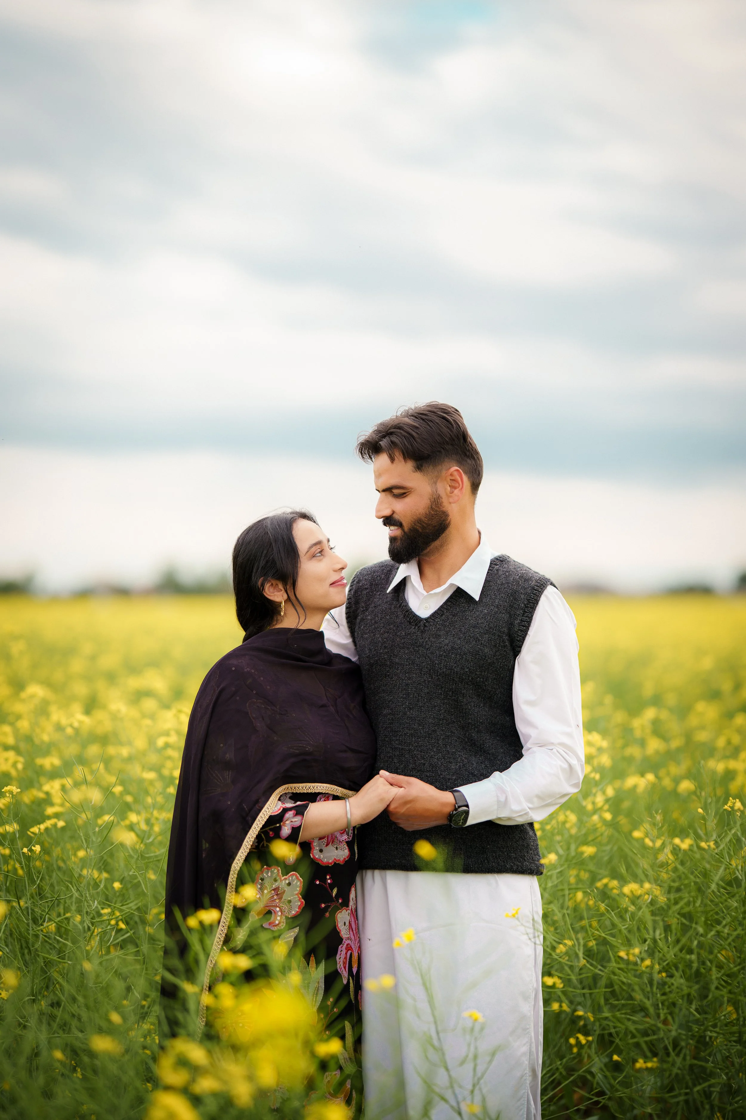 A couple standing close together in a yellow flower field, gazing into each other's eyes, with a cloudy sky above.