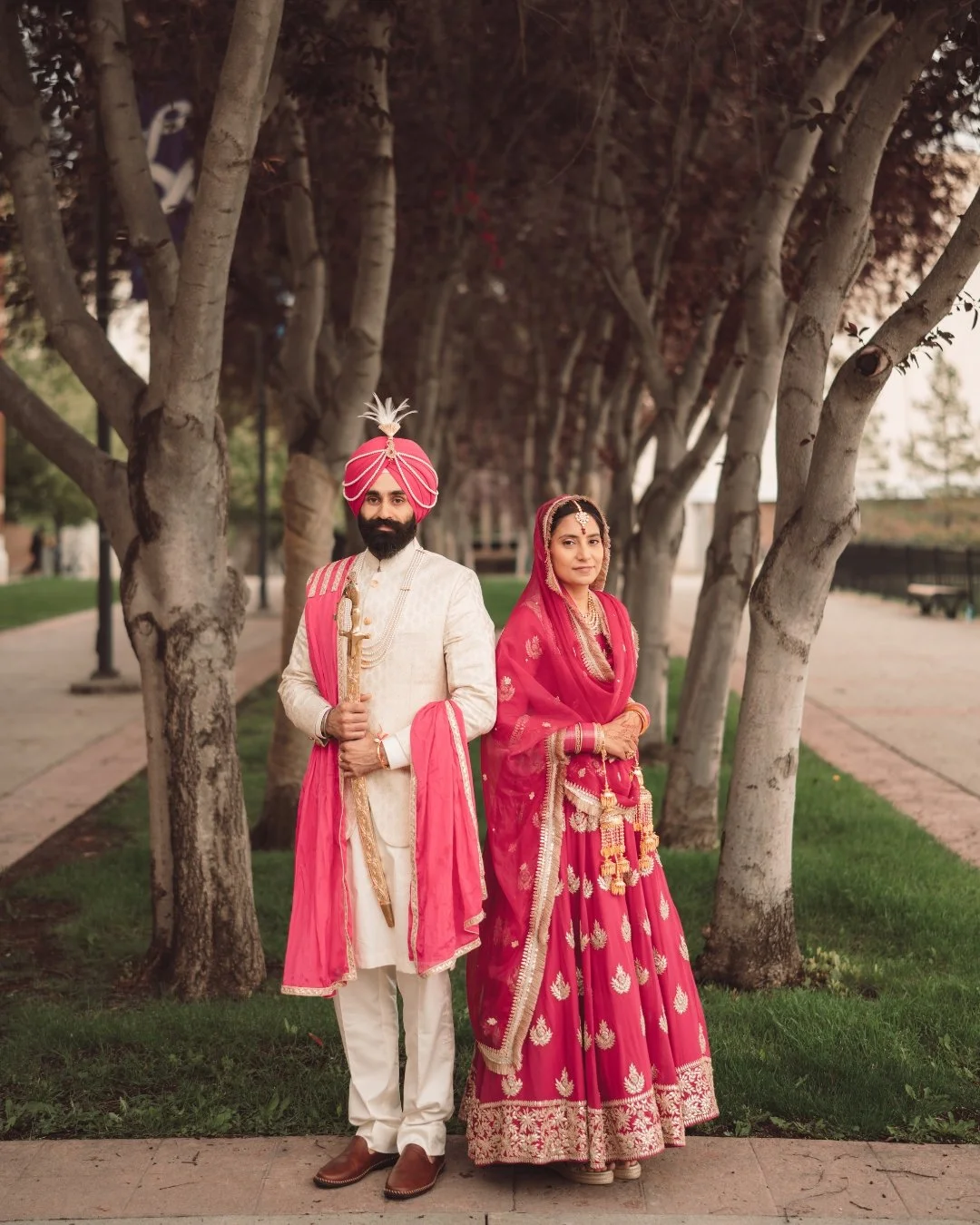 A man and woman in traditional Indian wedding attire standing outdoors under a row of trees.