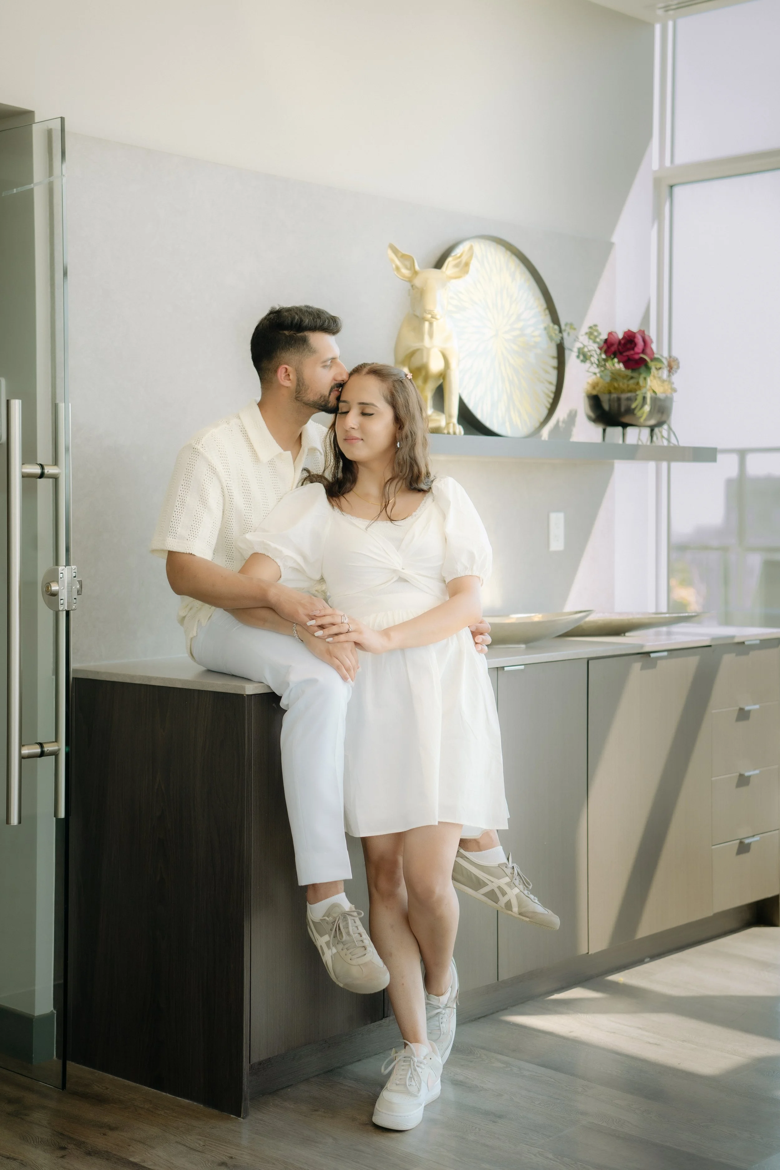 A couple dressed in white sits in a bright, modern kitchen. The man is kissing the woman's forehead while she has her eyes closed. They are leaning against a countertop with decorative items, including a gold sculpture of a deer and a black bowl with