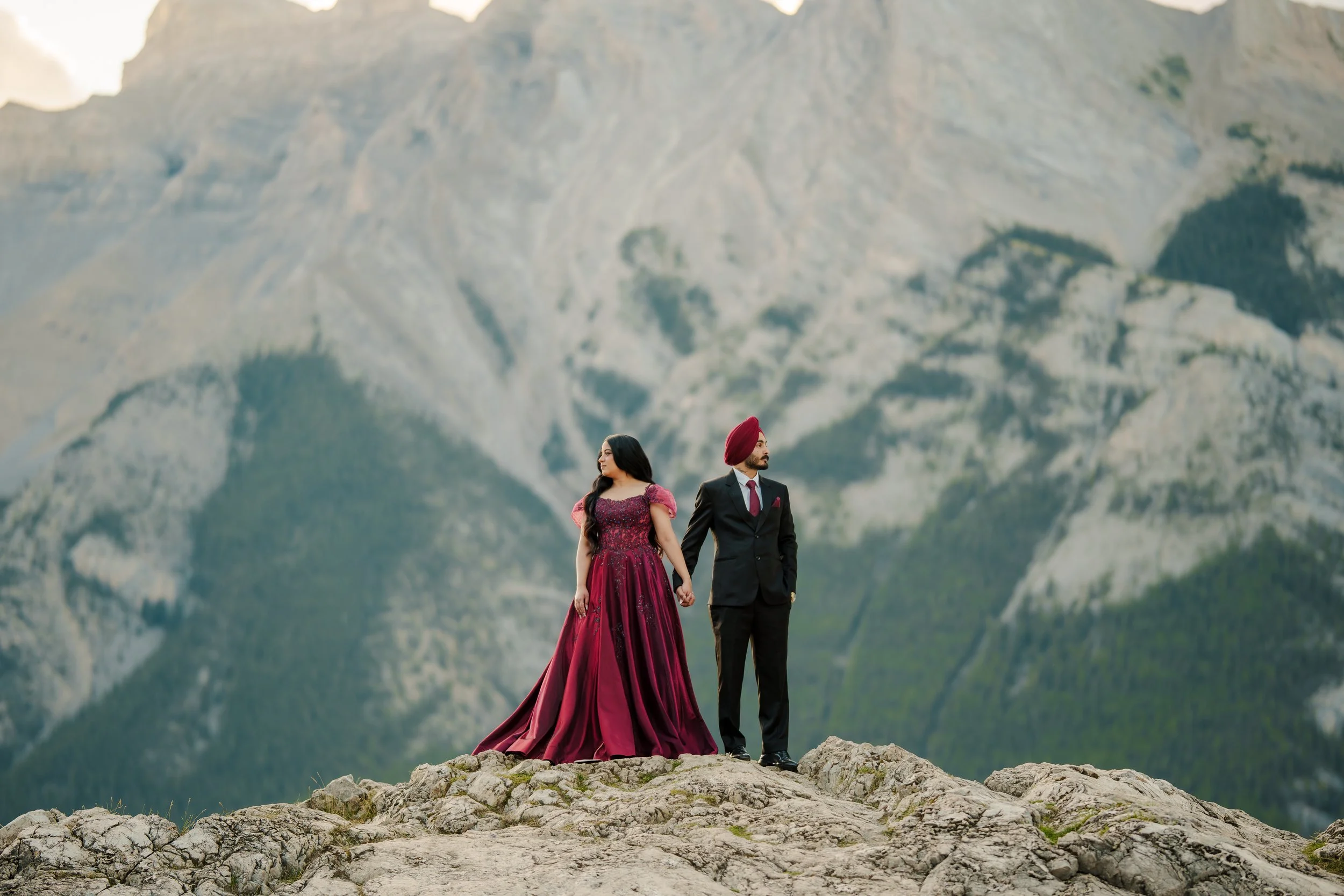 A woman in a long, elegant magenta dress and a man in a black suit with a red turban holding hands on a rocky outcrop with a mountain range in the background.