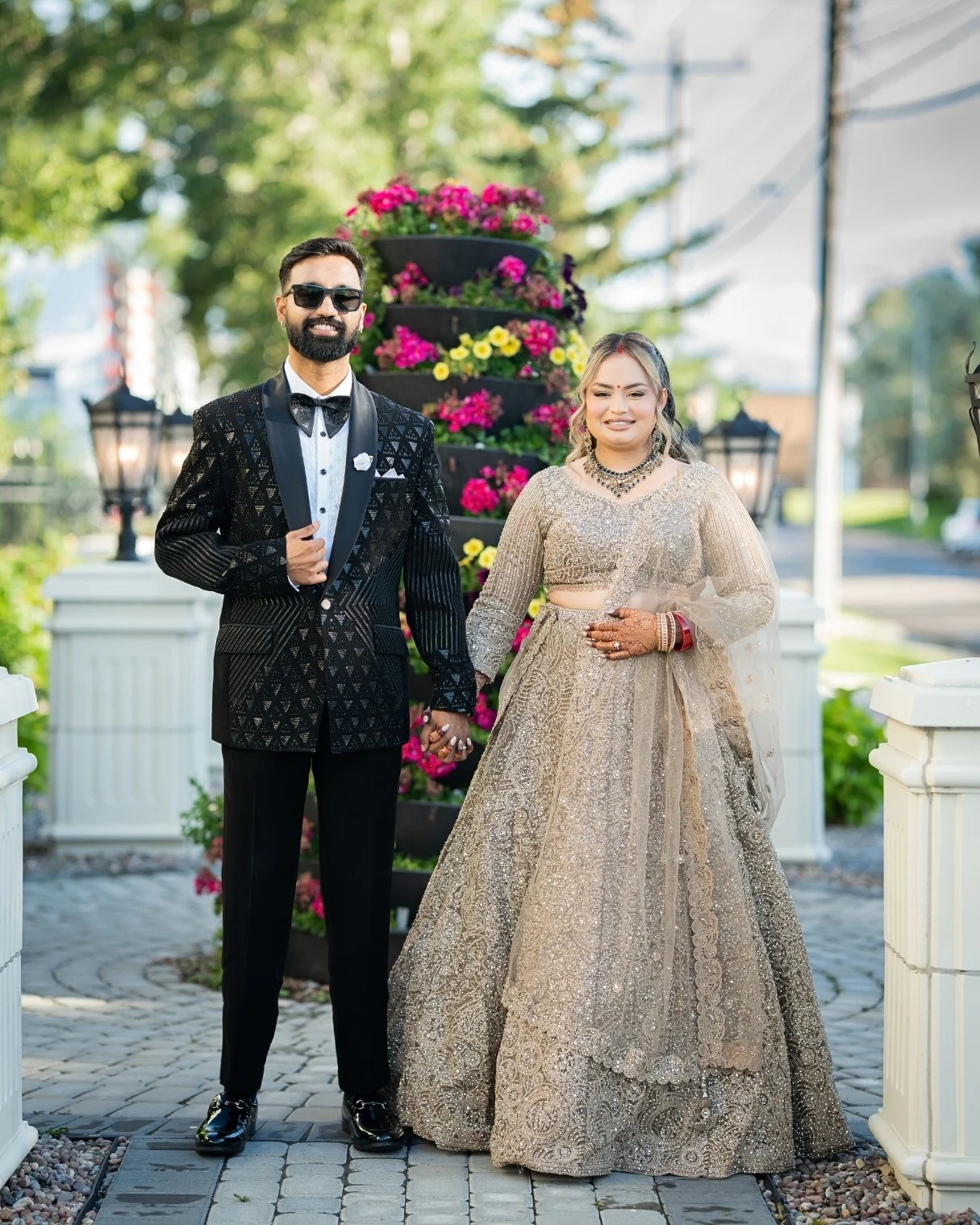 A smiling man in a black tuxedo with a bow tie and sunglasses, holding hands with a woman in a gold traditional dress and jewelry, standing outdoors with a floral arrangement and lanterns in the background.