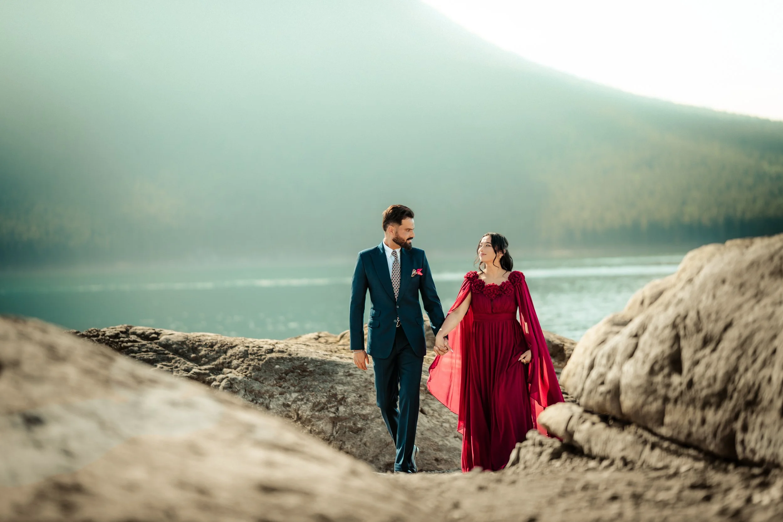 A man in a suit and a woman in a long red dress holding hands and walking on a rocky shoreline near a body of water with mountains in the background.