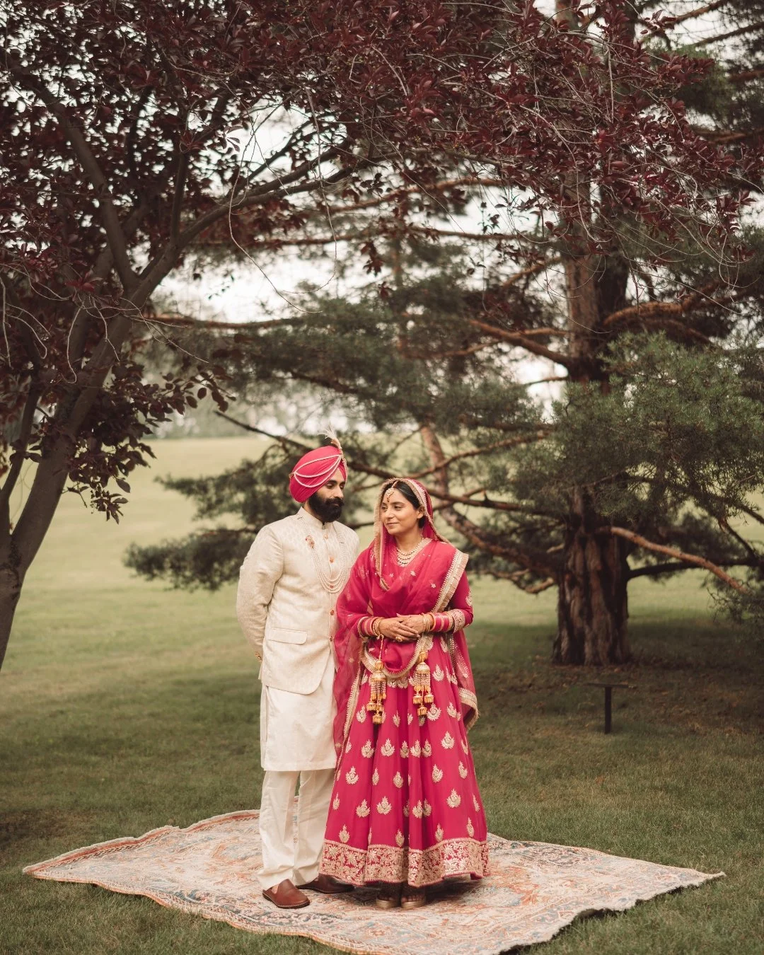 Indian couple in traditional wedding attire standing on a rug outdoors, surrounded by trees in a park.