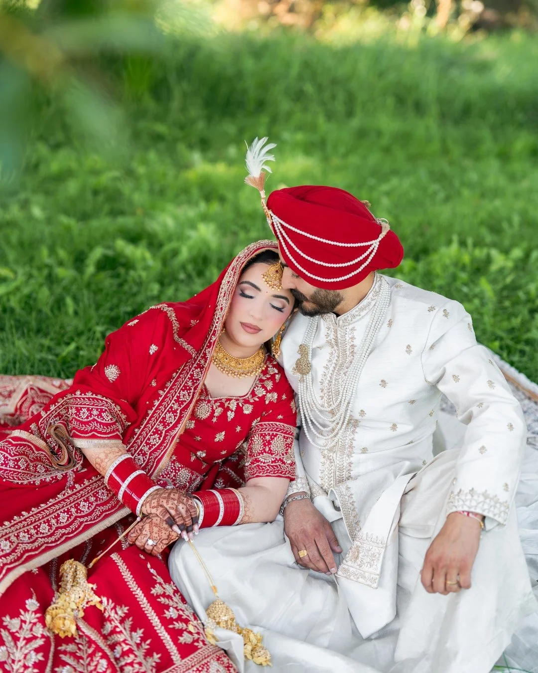 An Indian bride and groom sitting close with their foreheads touching, dressed in traditional wedding attire, outdoors on green grass.
