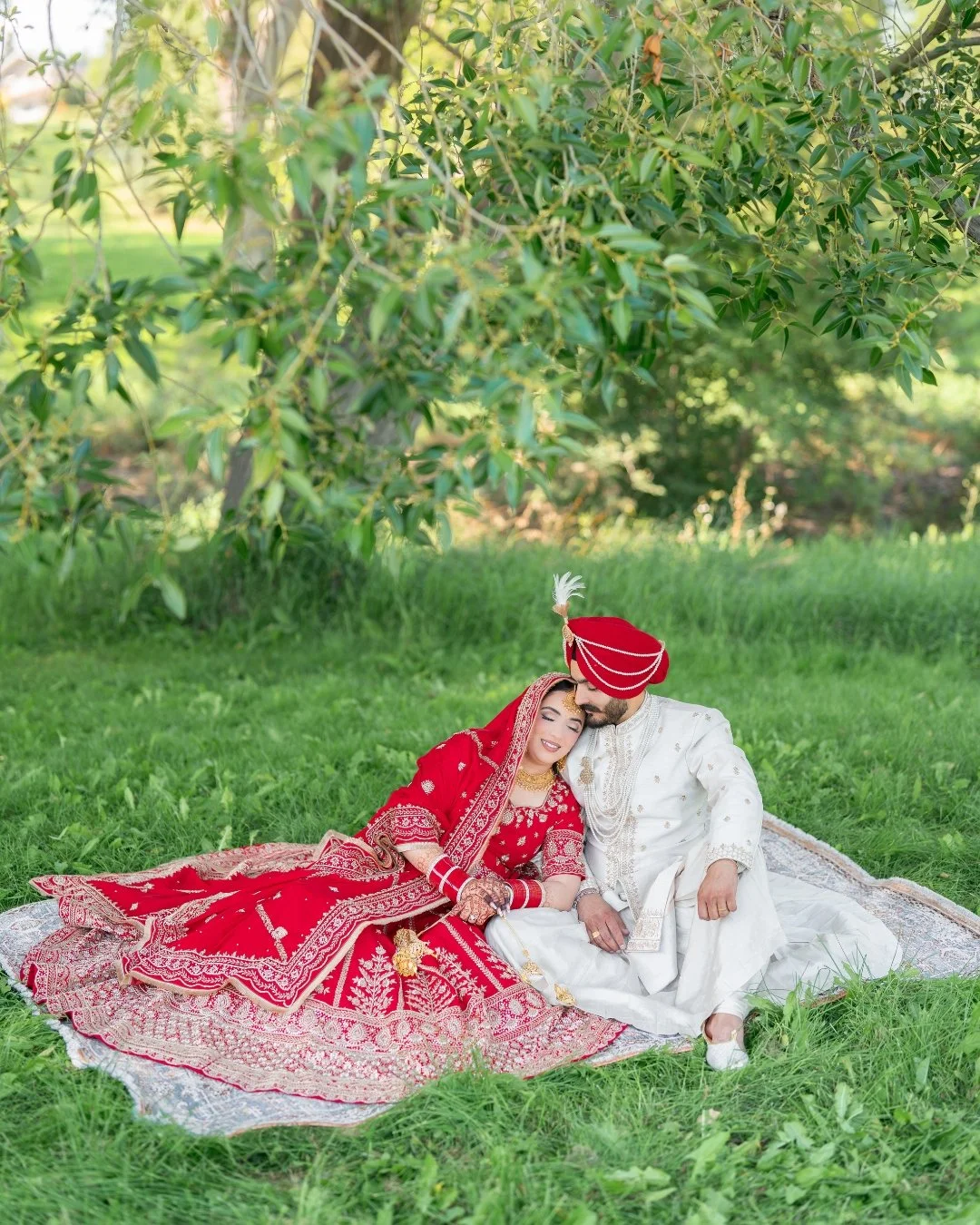 A newlywed couple dressed in traditional Indian wedding attire sitting on a blanket on the grass under a tree, with the bride resting her head on the groom's shoulder and both smiling.