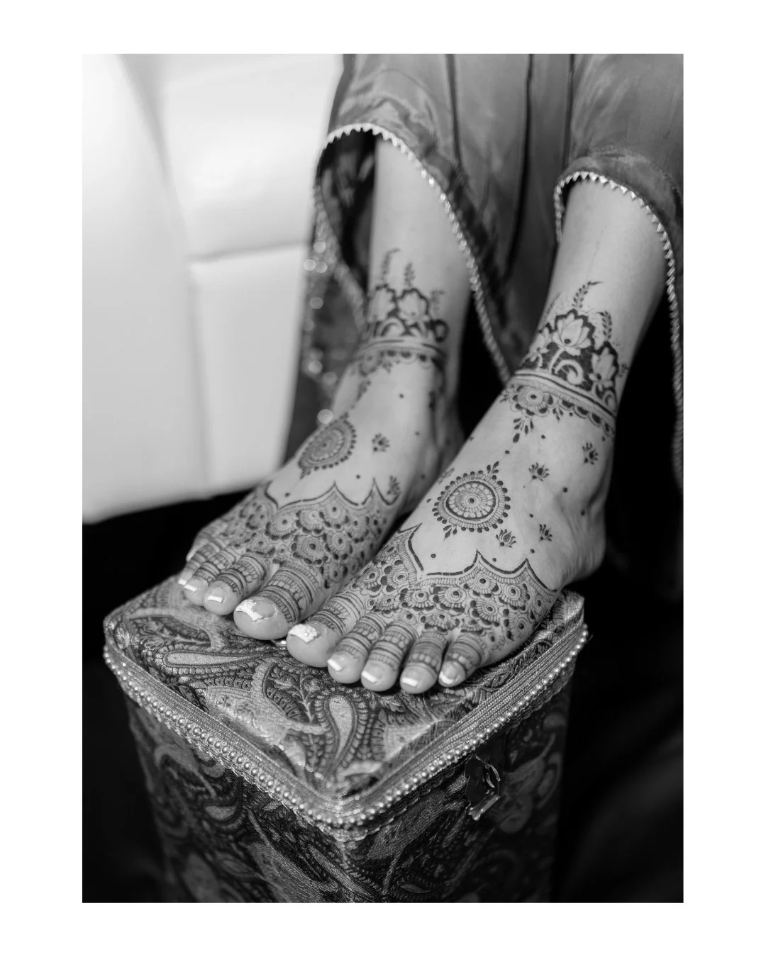 Black and white photo of heavily henna-decorated hands resting on ornate fabric box, with intricate floral and geometric designs on fingers and wrists. Mendi day