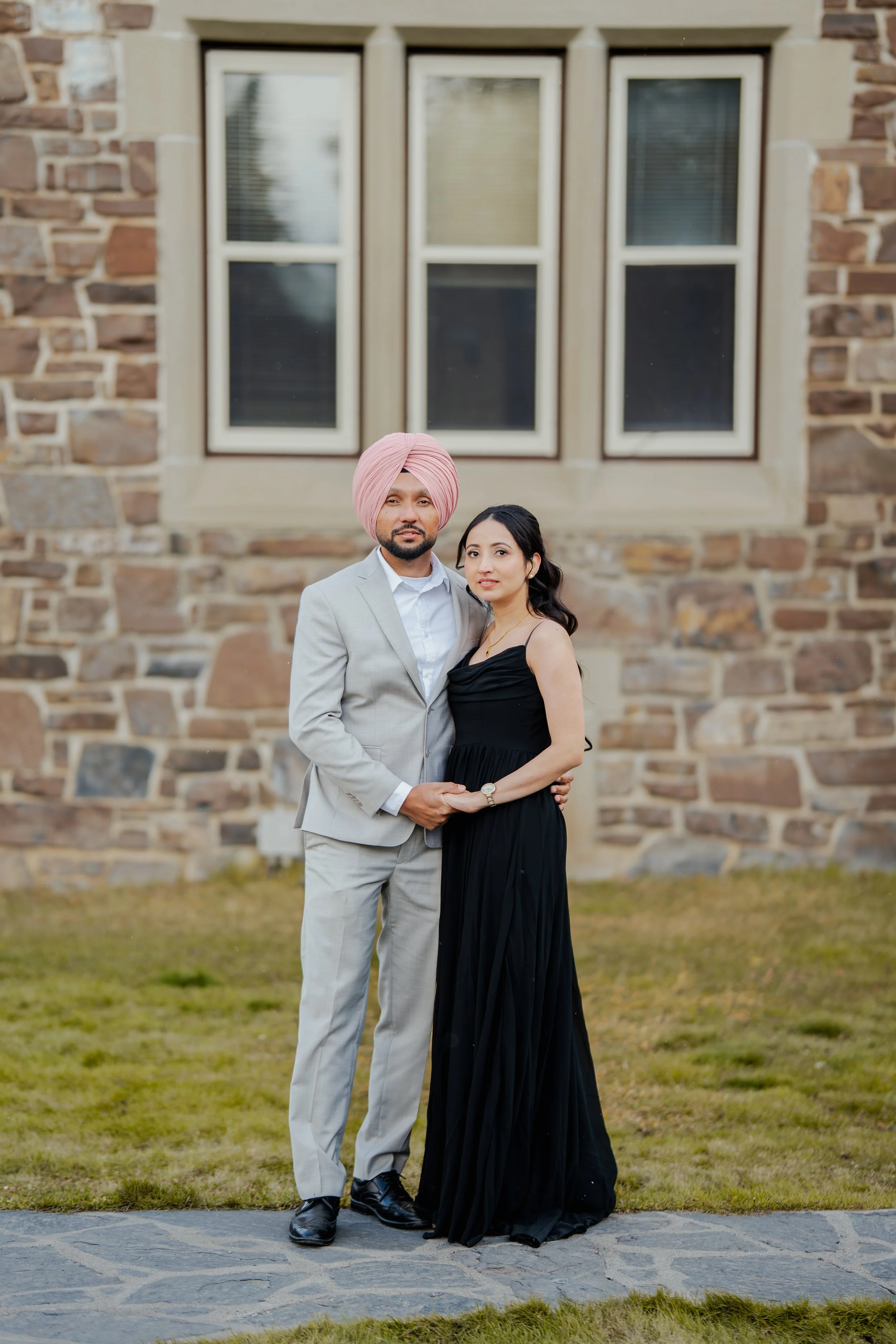 A couple standing outdoors, the man wearing a light grey suit and a pink turban, the woman wearing a black dress, in front of a brick building with large windows. E-shoot photos
