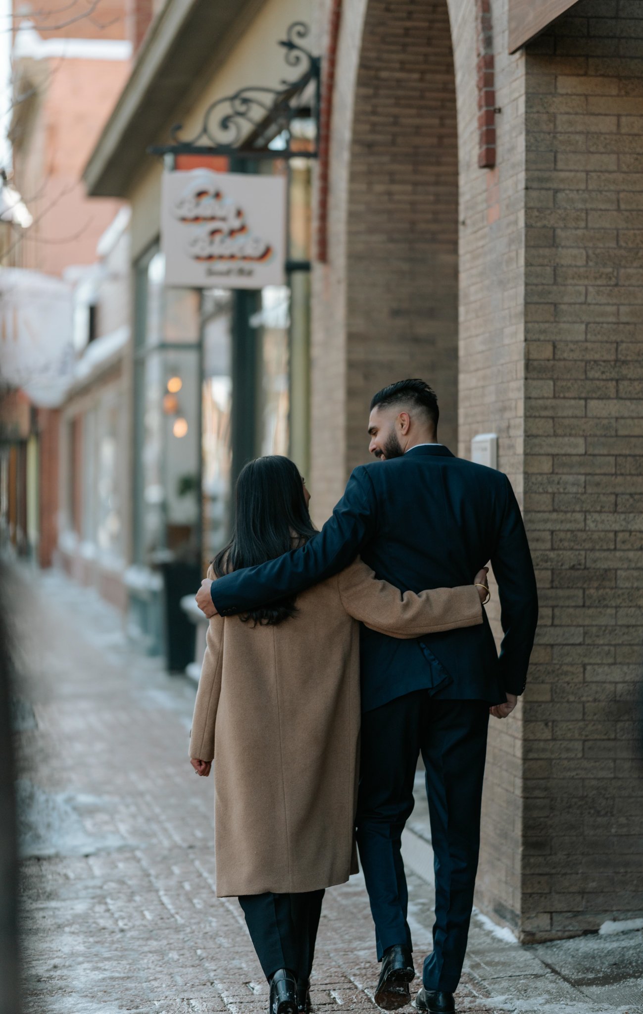 A man and woman walking closely together along a brick sidewalk, with the man’s arm around the woman’s shoulders, engaged in conversation.
