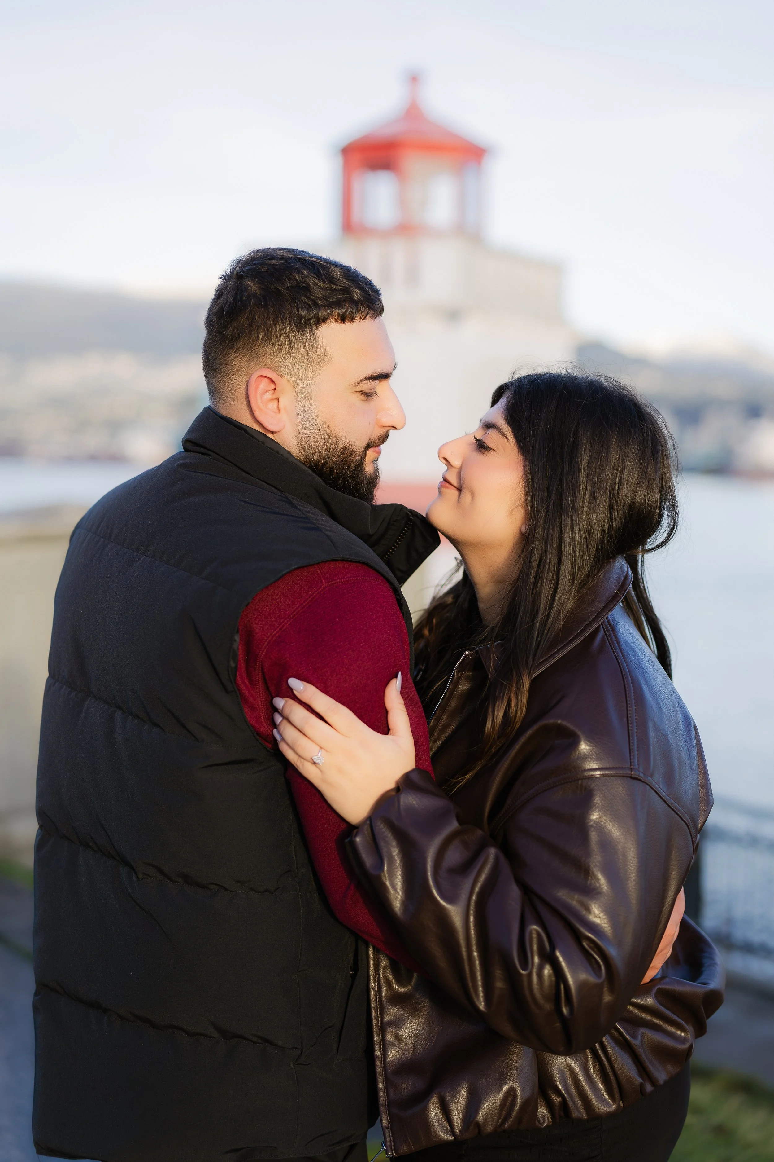 A couple embracing outdoors, with a lighthouse in the background, during daytime.