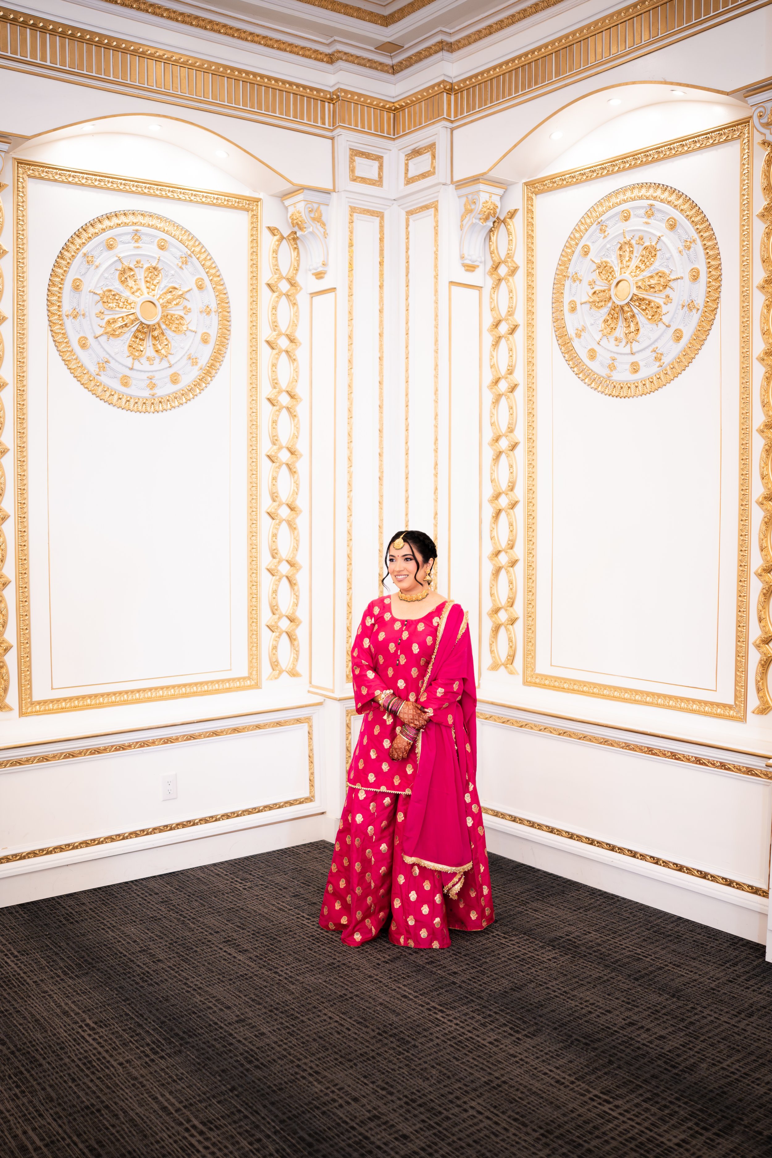 A woman in a pink traditional Indian dress with gold accents, standing in a room with ornate white and gold walls, smiling.